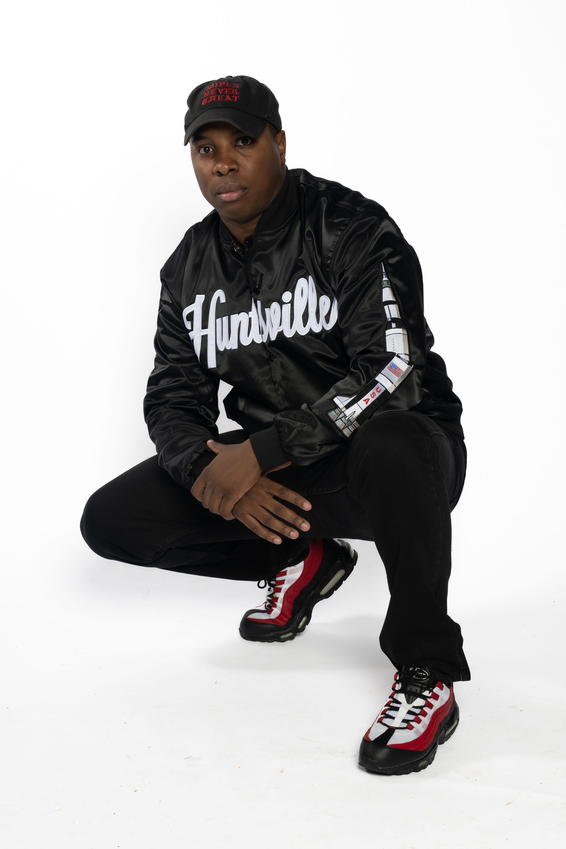 Person crouching in a black jacket, cap, and red-accented sneakers against a white background