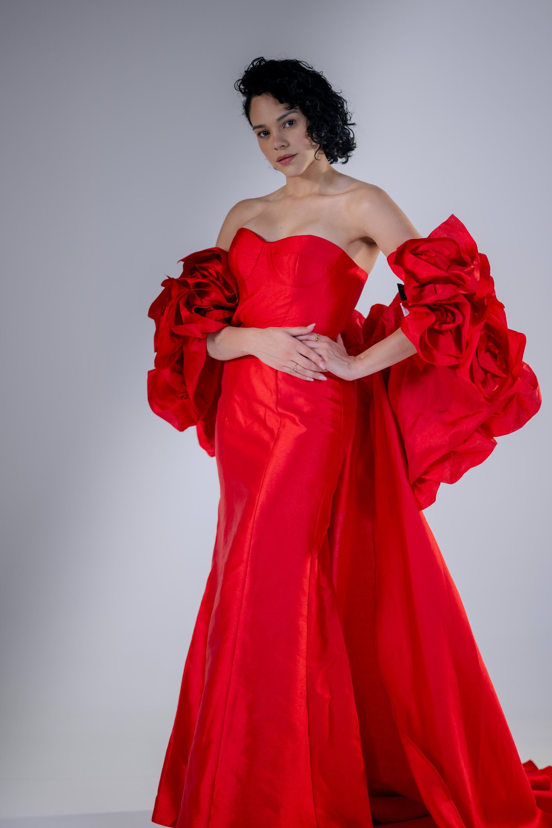 Woman in a bright red off-shoulder gown posing against a plain gray backdrop.
