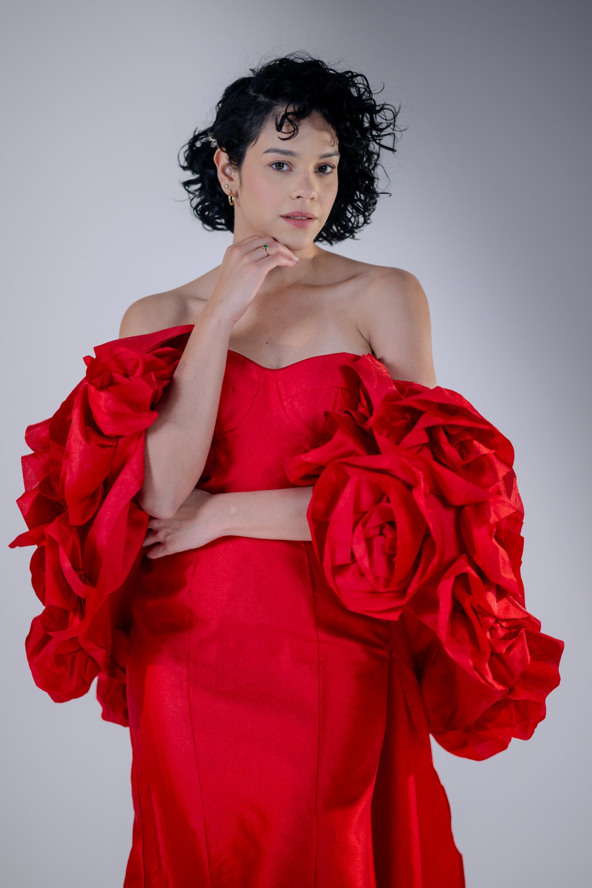 Person in a bright red off-shoulder dress with large ruffled sleeves, posing against a gray studio backdrop
