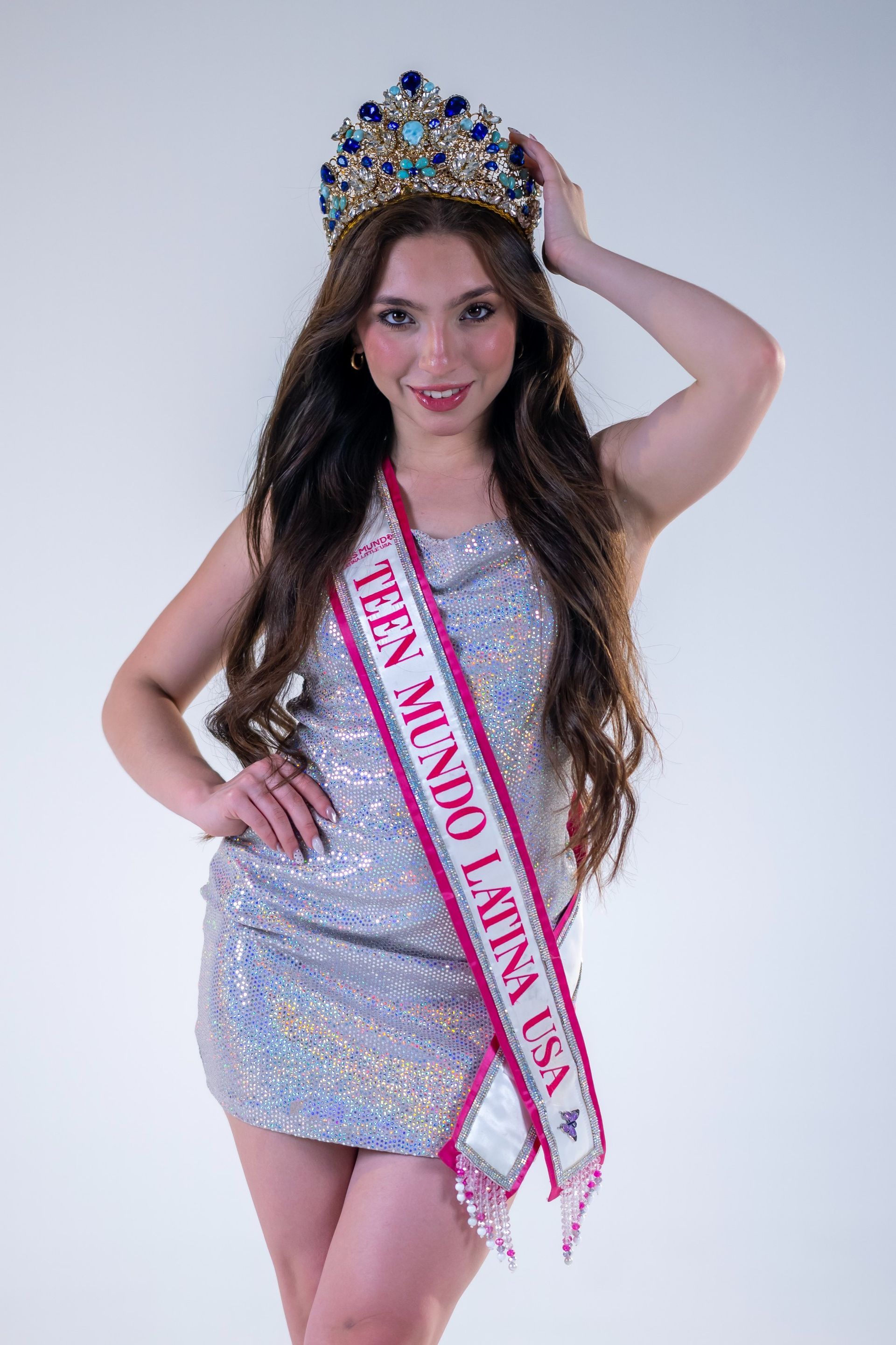 Woman in a sparkly silver dress and crown, wearing a pageant sash against a white background.