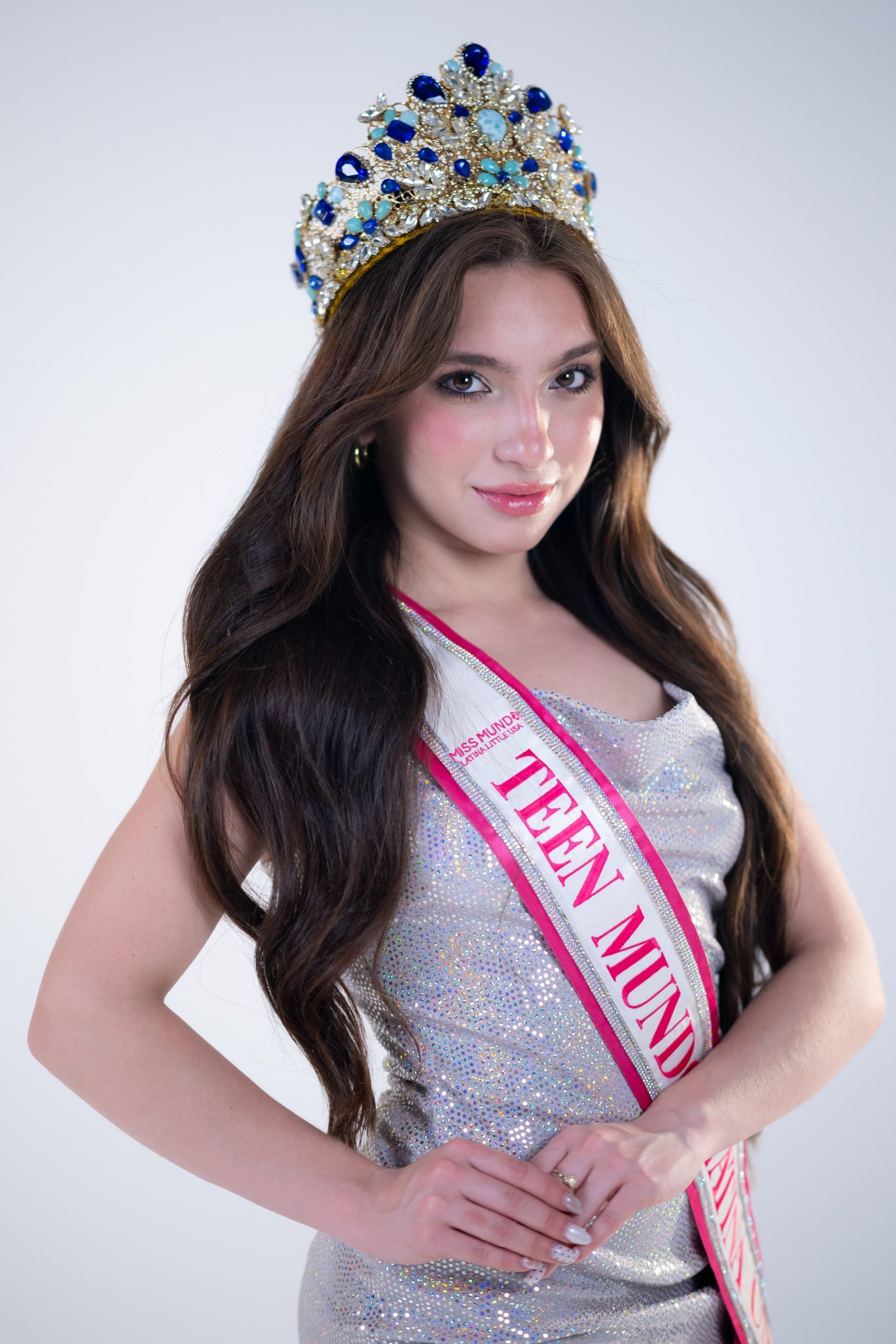 Woman wearing a jeweled crown and pink sash, posing against a light background.