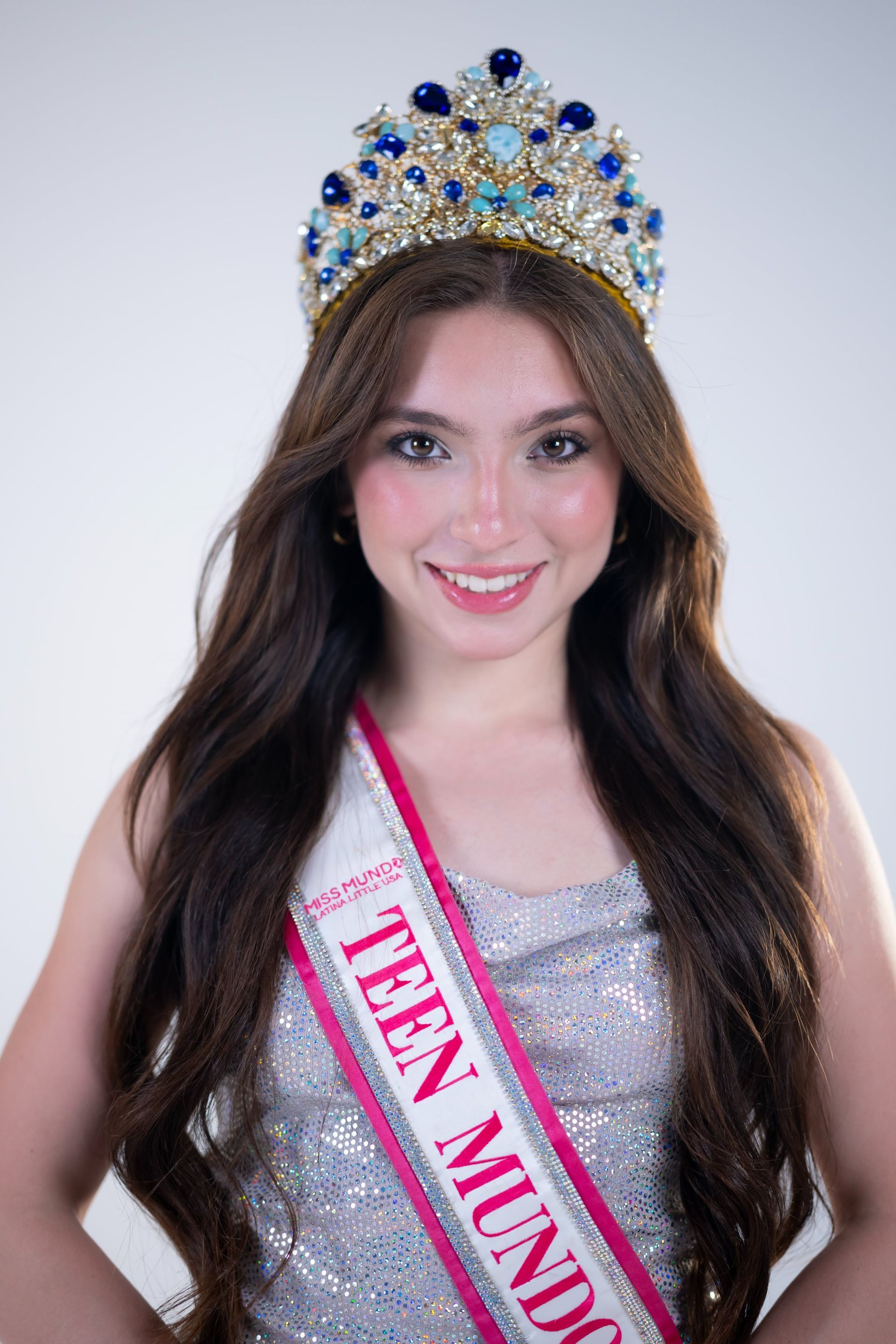 Young woman in jeweled crown and pink sash, smiling against a white background