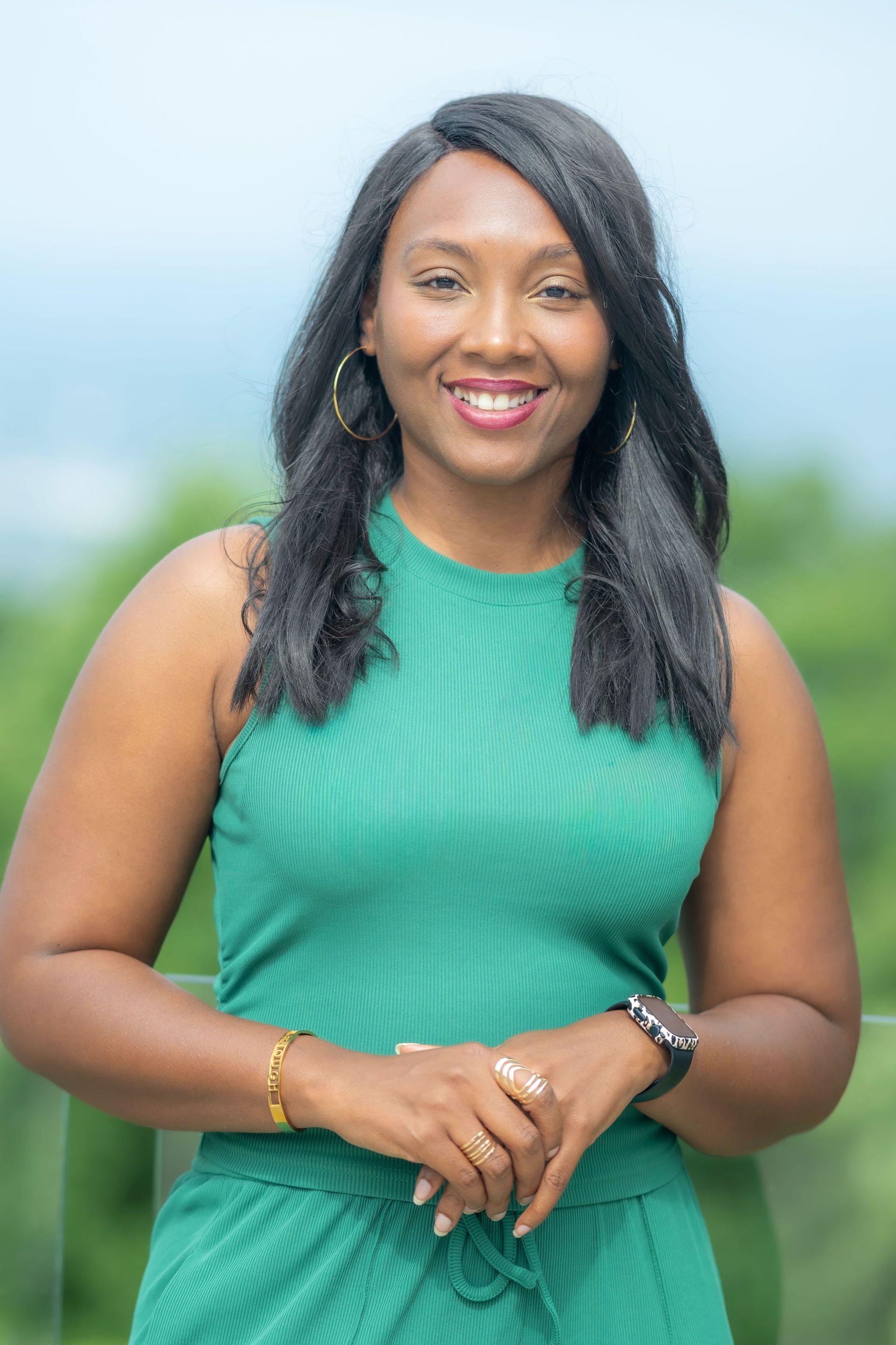 Woman in a green sleeveless top smiling outdoors with a blurred blue-green background