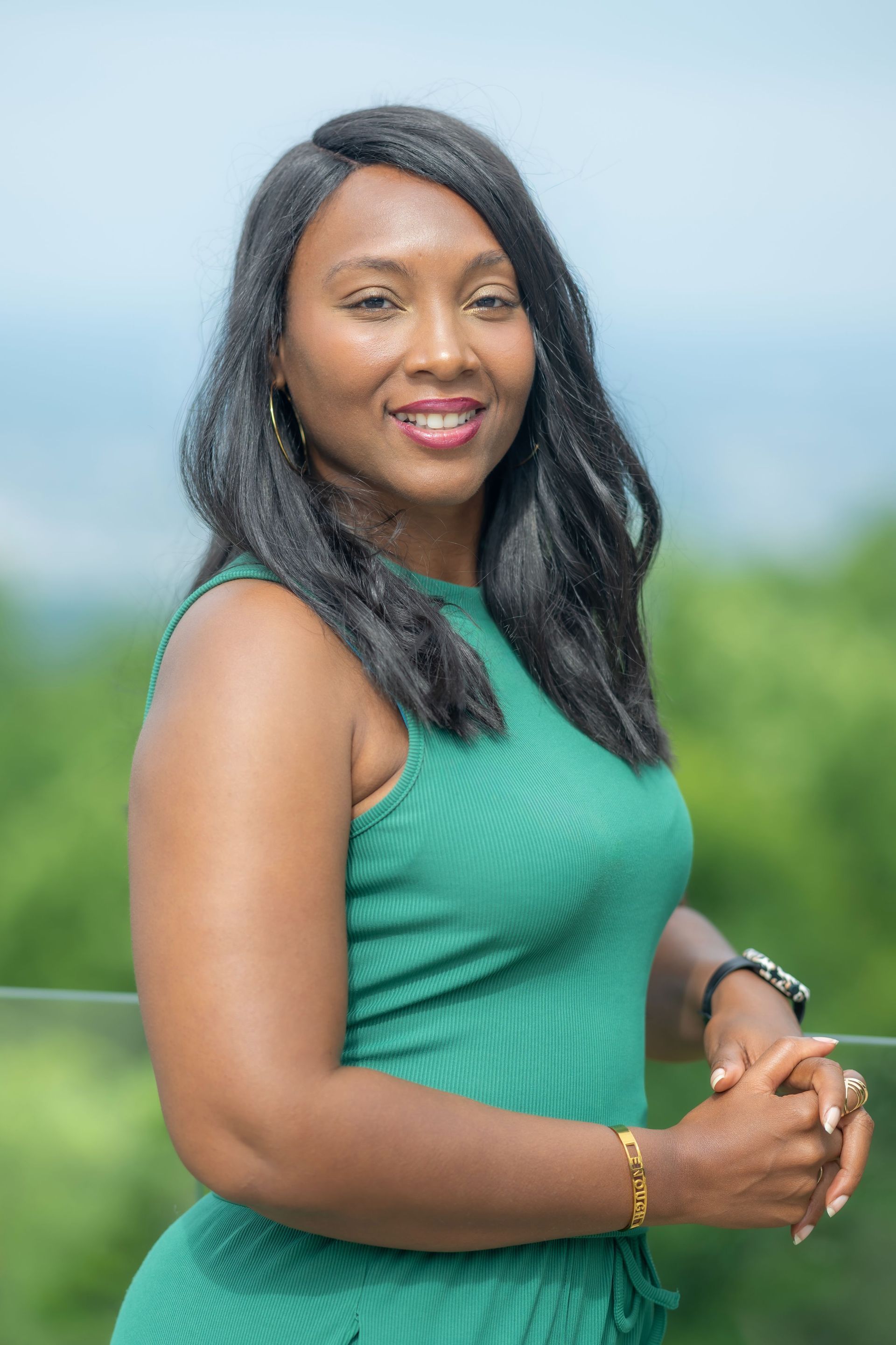 Woman in a green sleeveless dress standing outdoors with a blurred green landscape behind her.