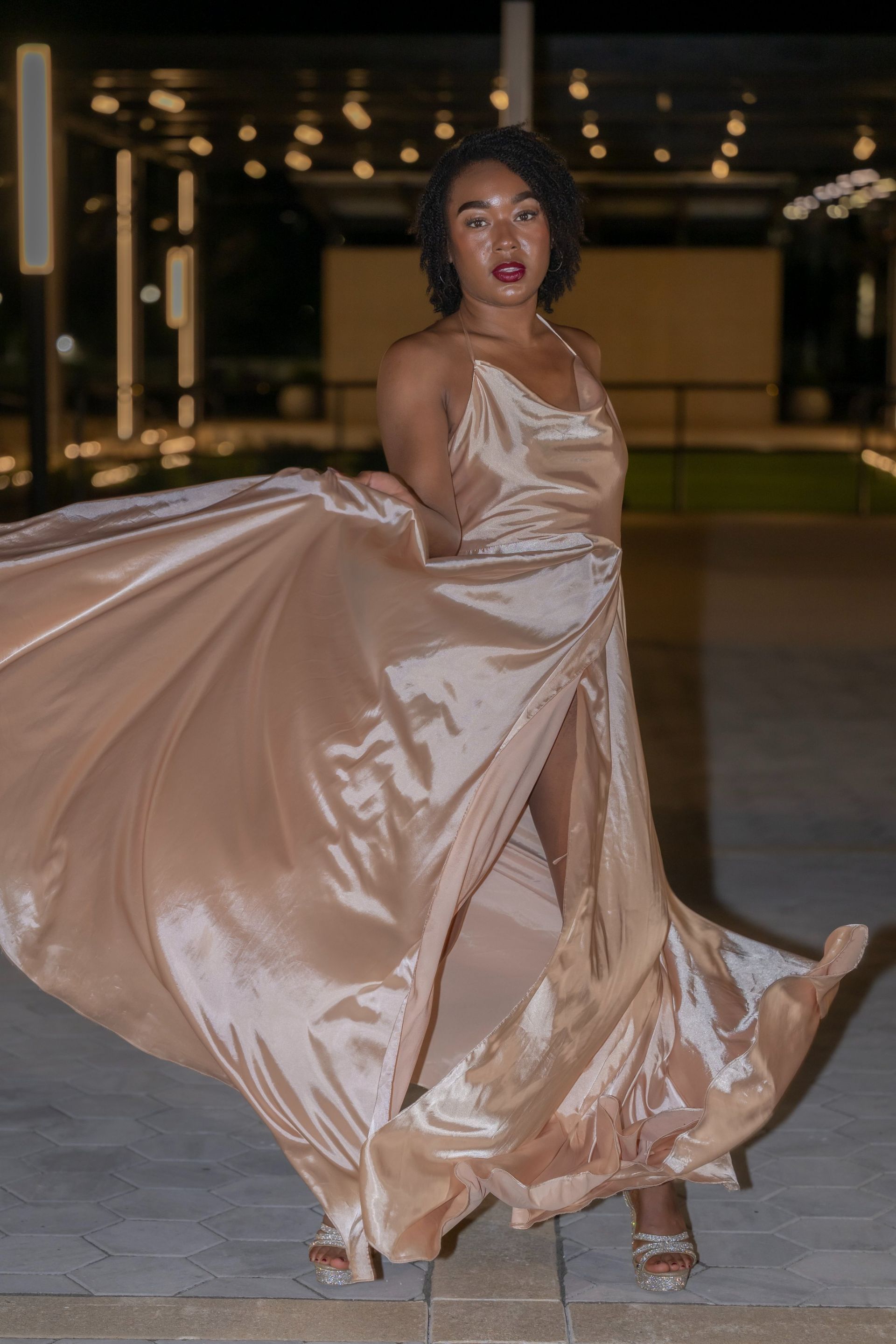 Woman in a flowing champagne satin gown twirls on a nighttime walkway, dress billowing wide.