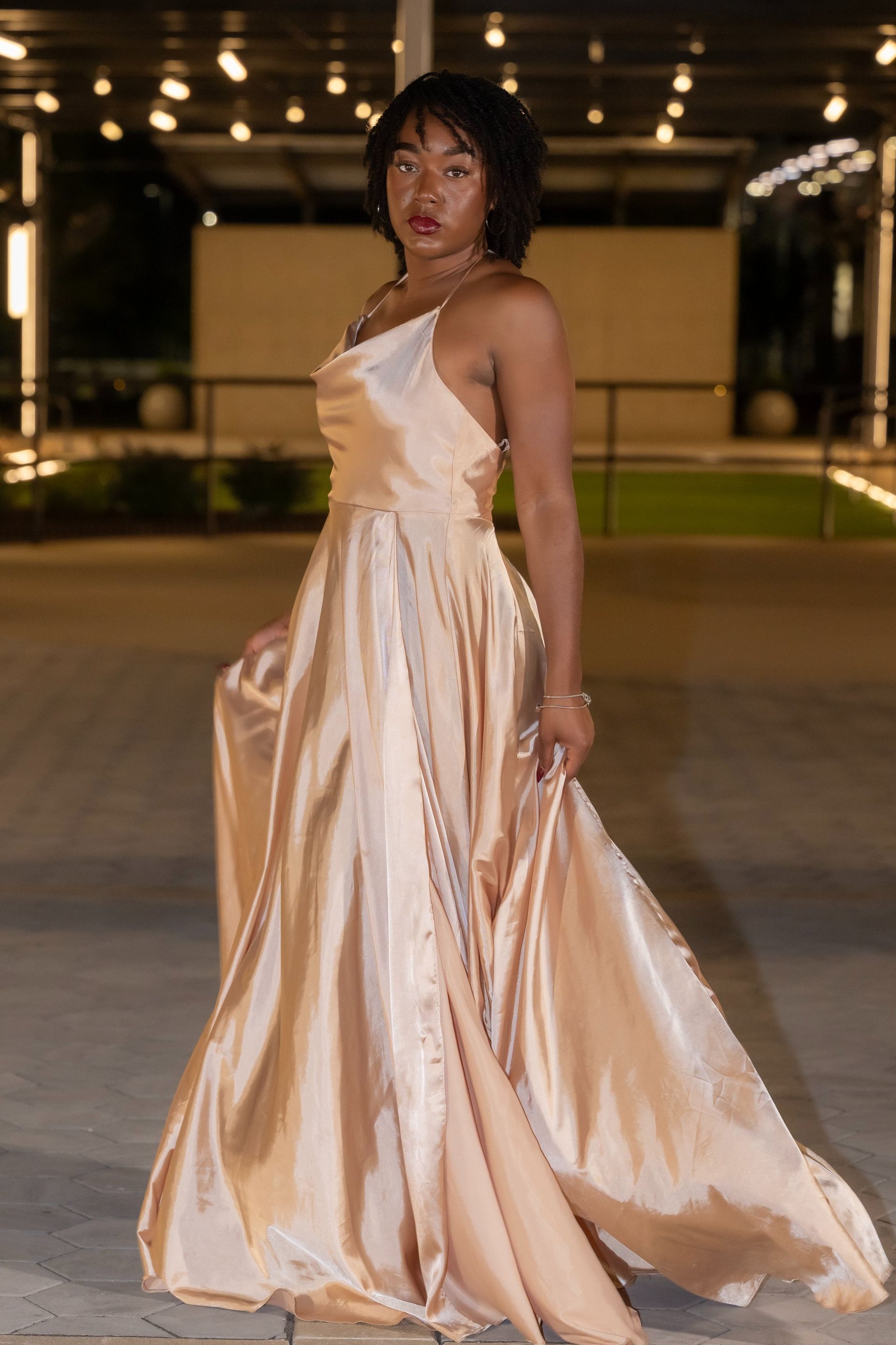 Person in a champagne satin gown posing outdoors at night under warm string lights.