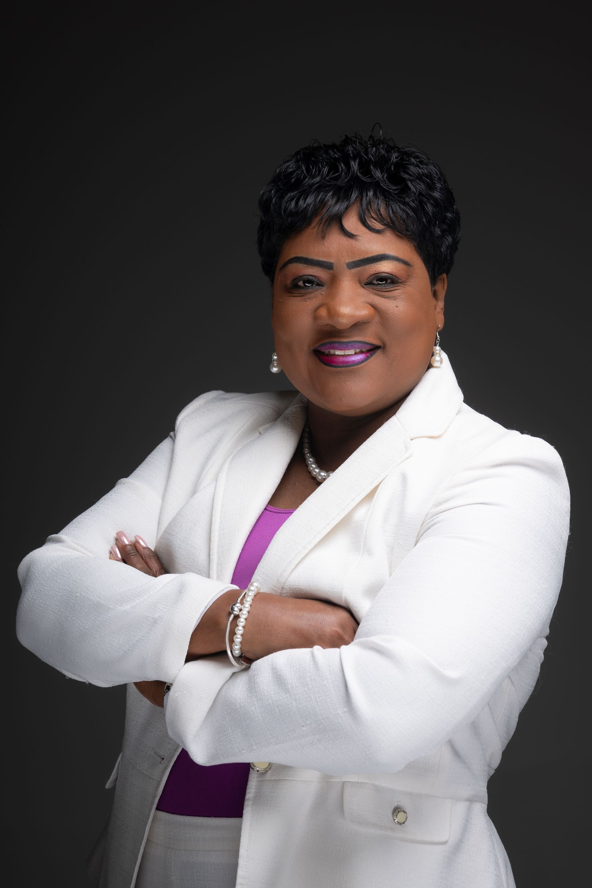 Smiling professional in a white blazer and purple top, arms crossed against a dark background