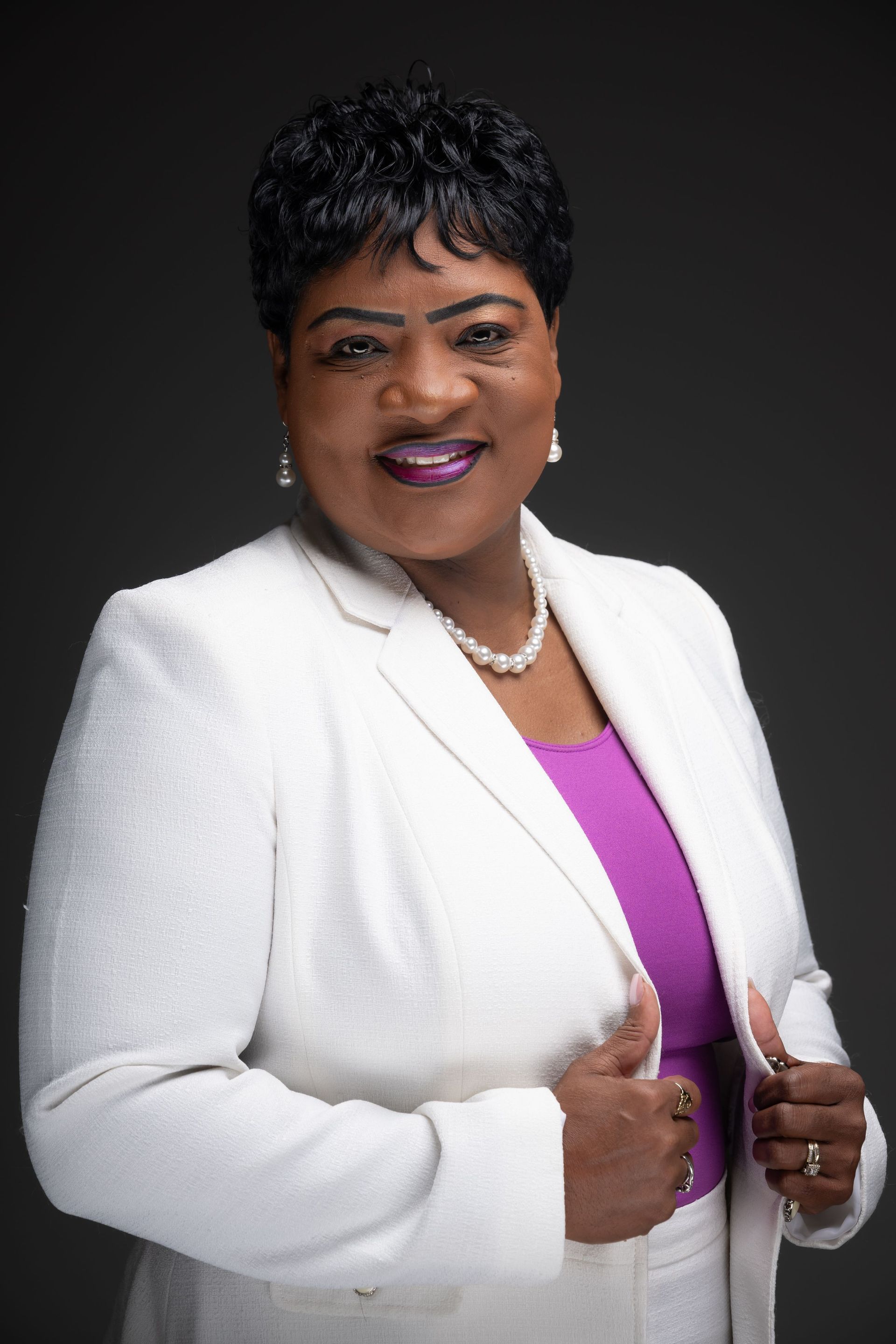 Studio portrait of a smiling woman in a white blazer over a purple top, against a dark background