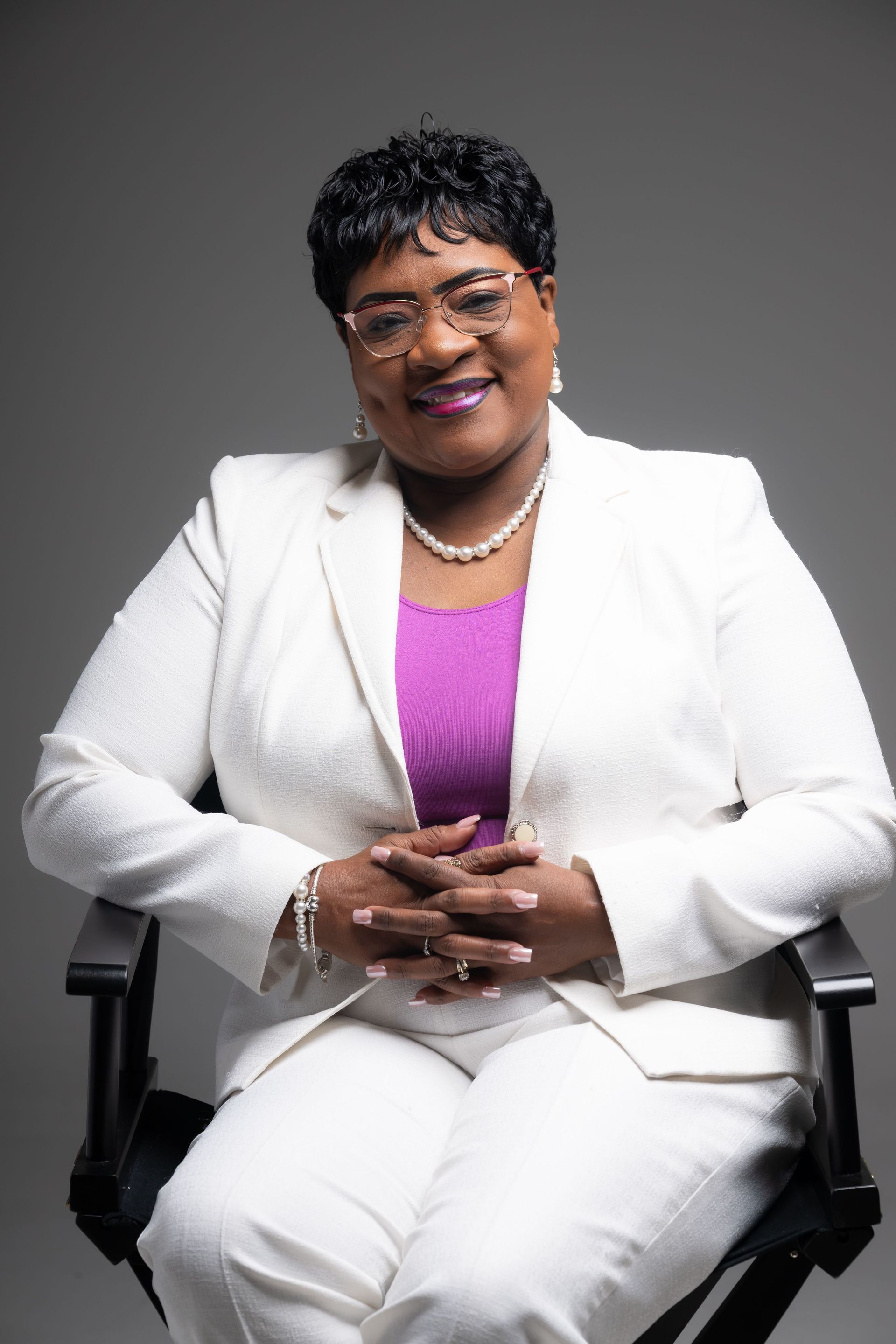 Smiling person in a white suit and purple top seated against a gray backdrop