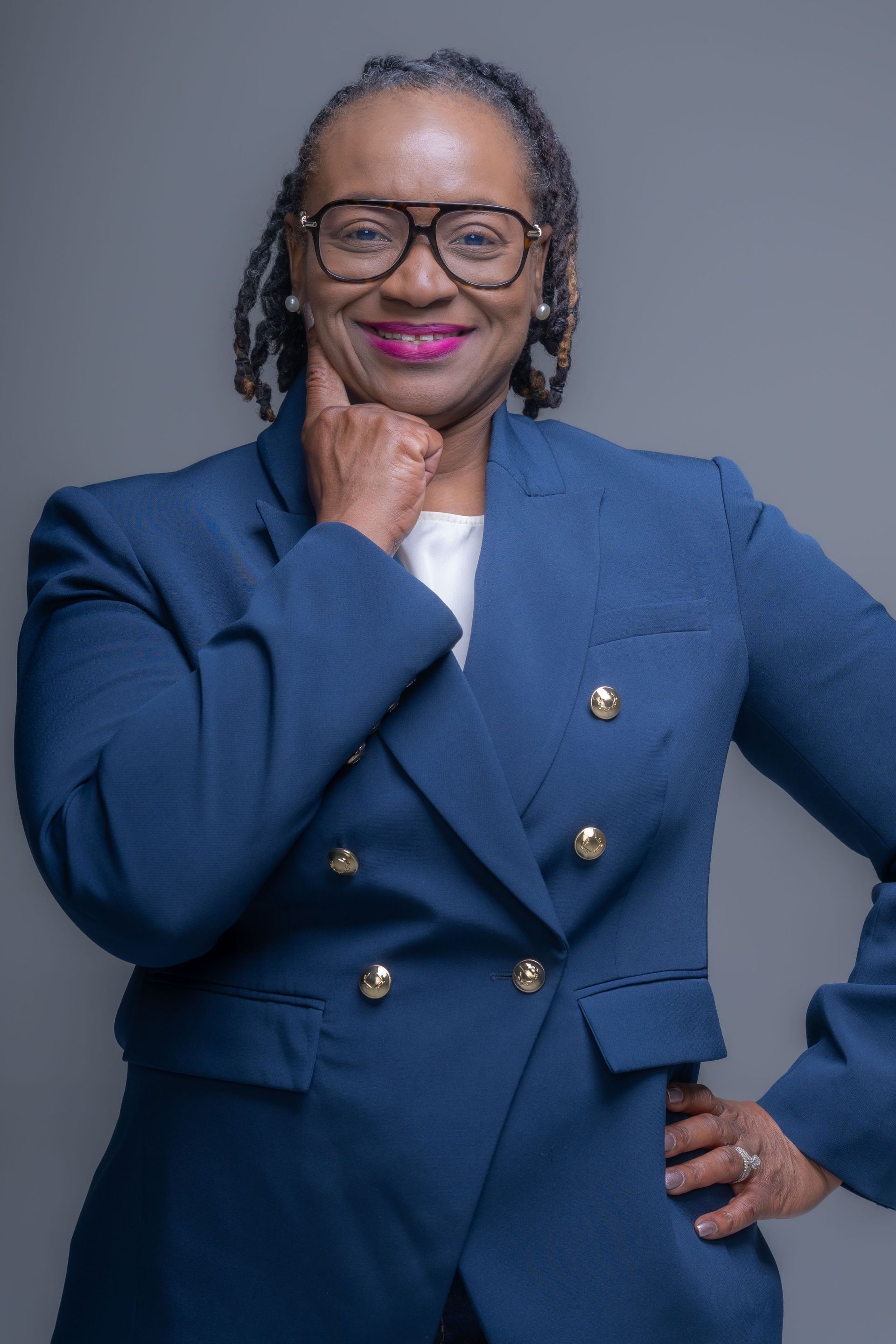 Smiling person in a blue blazer, glasses, and pink lipstick posing against a gray background