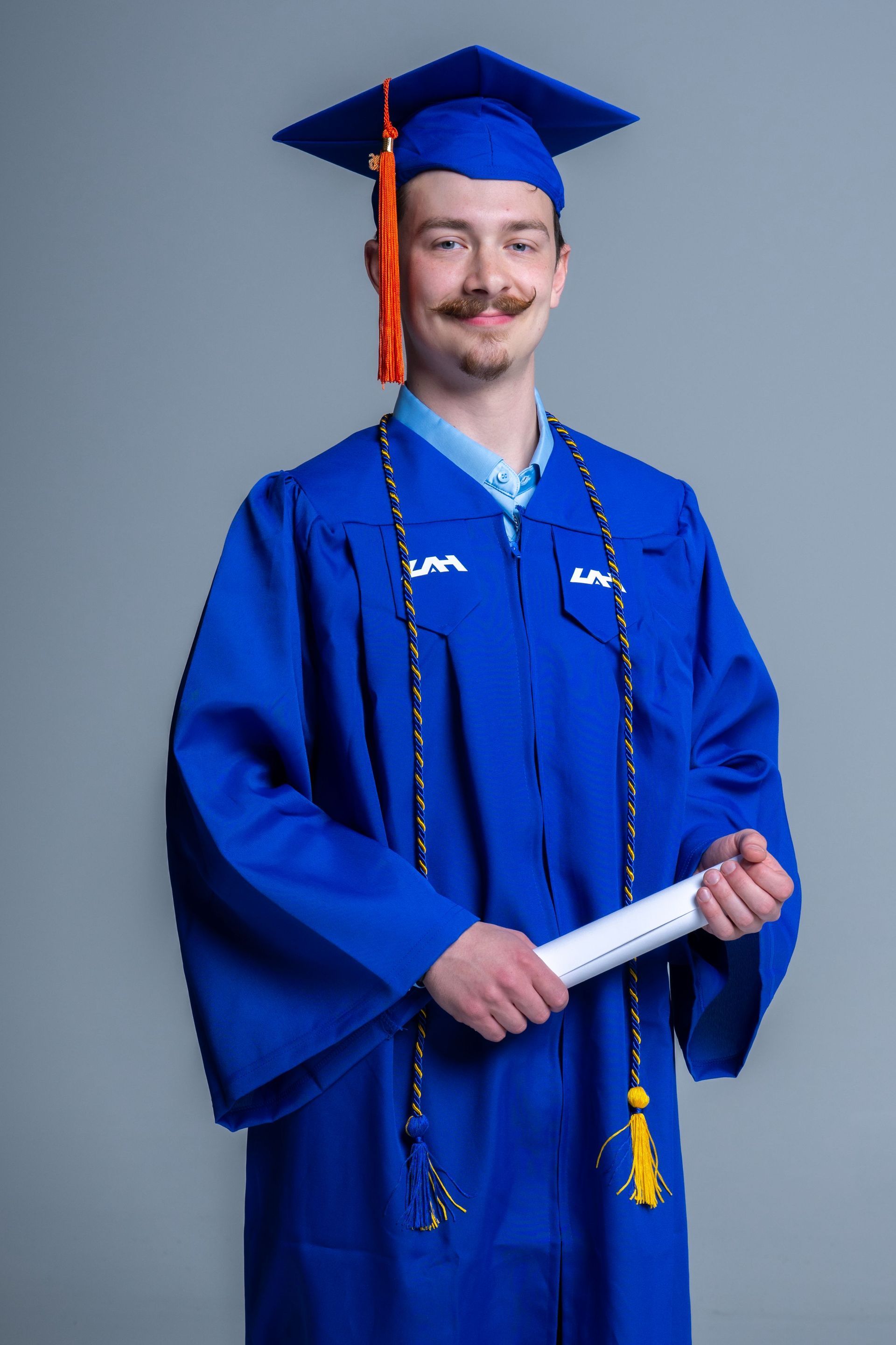 Graduate in blue cap and gown holding a diploma, smiling against a gray background