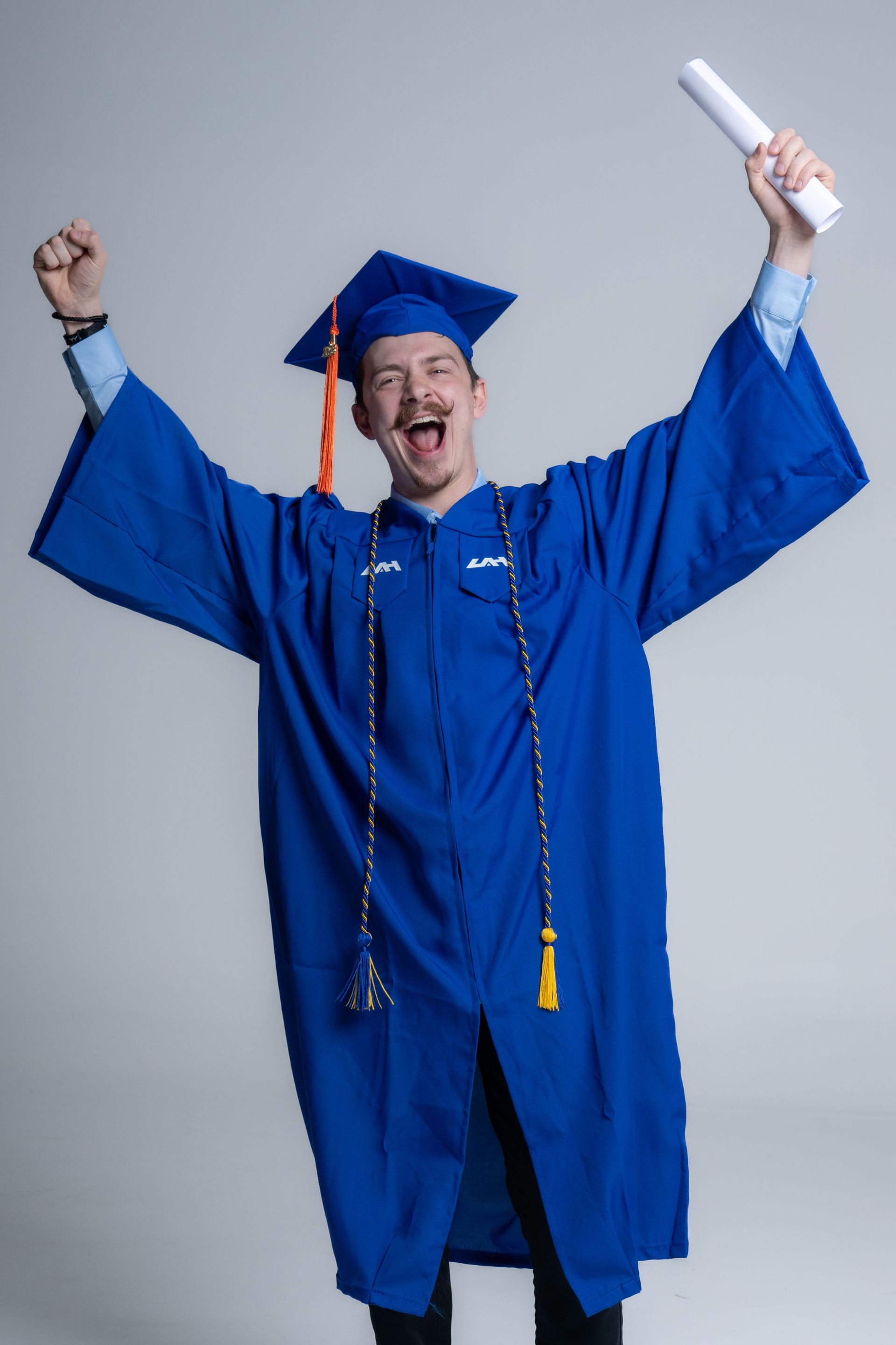 Smiling graduate in blue cap and gown holding diploma aloft with raised arms