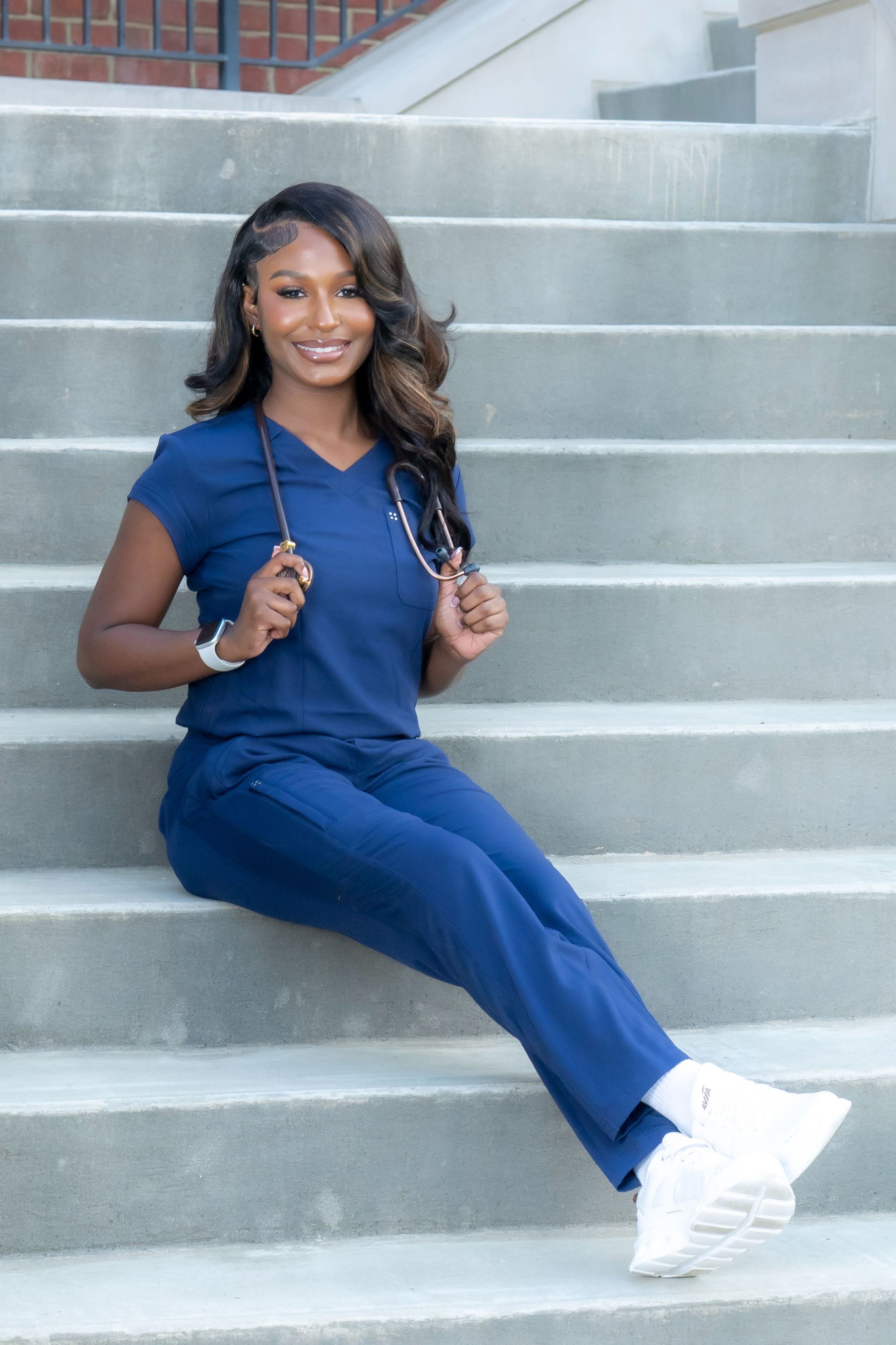 Person in blue scrubs sitting on outdoor concrete steps, smiling and holding stethoscopes.
