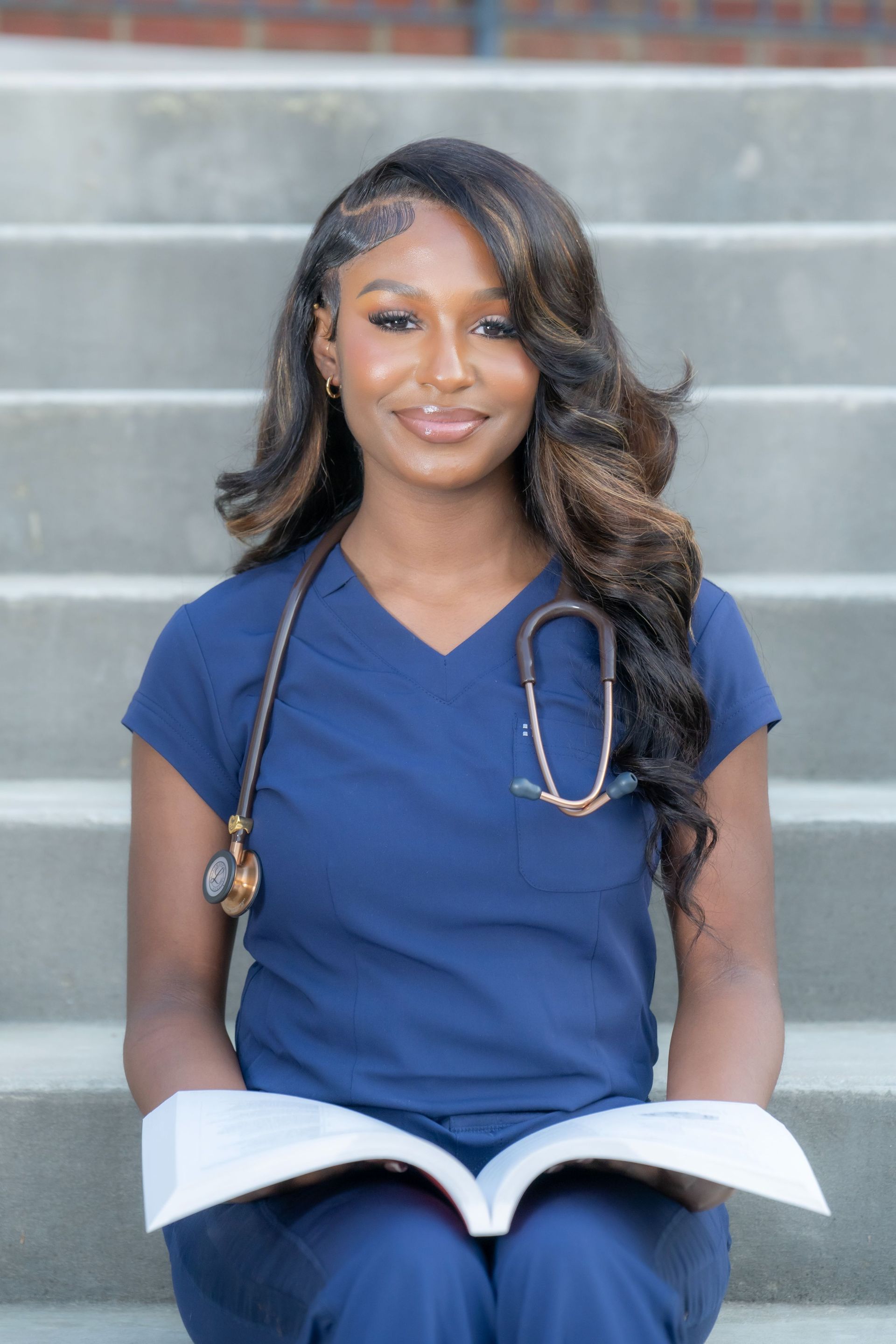 Smiling nurse in blue scrubs with a stethoscope, seated on steps reading an open book.