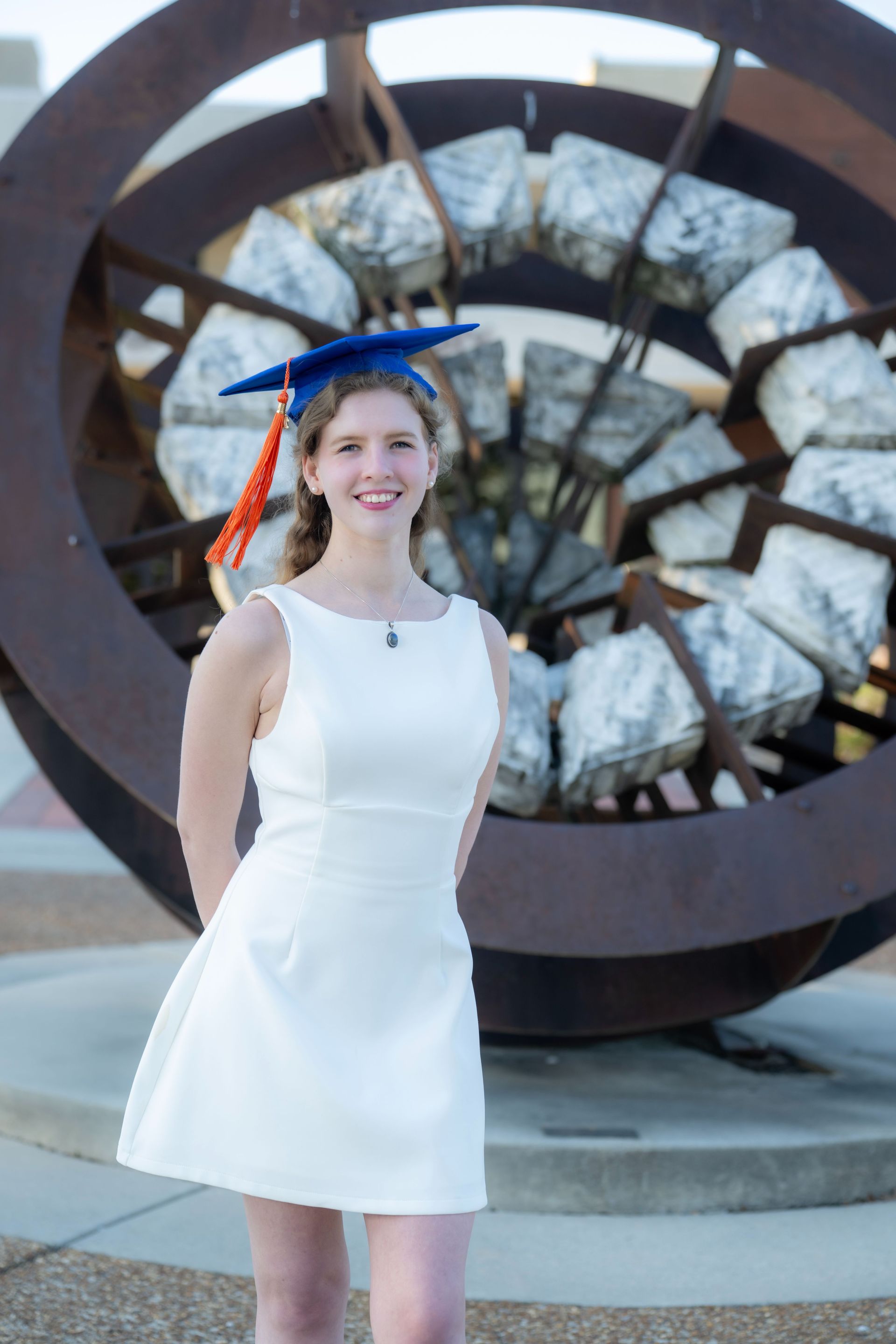 Woman in a white dress and blue cap standing before a large sculpture outdoors