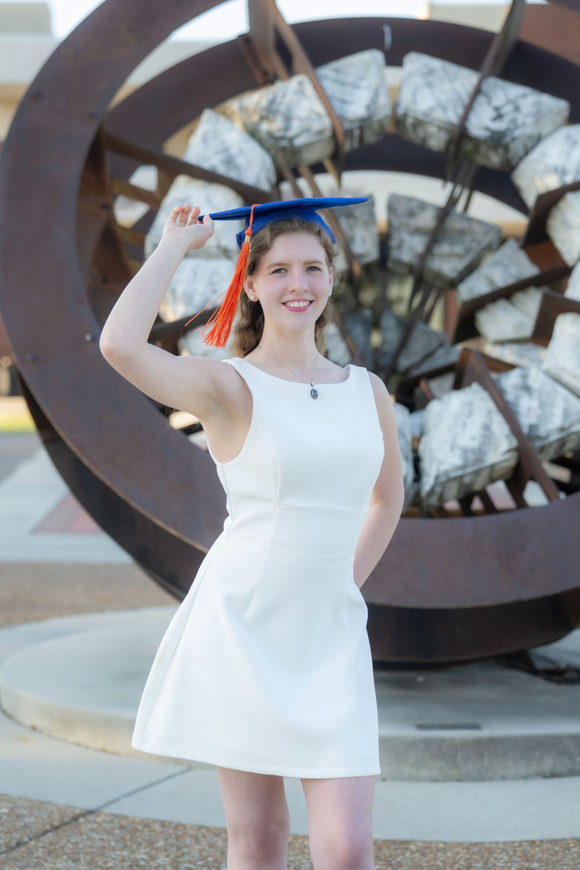 Woman in a white dress posing outdoors in front of a large circular metal sculpture.