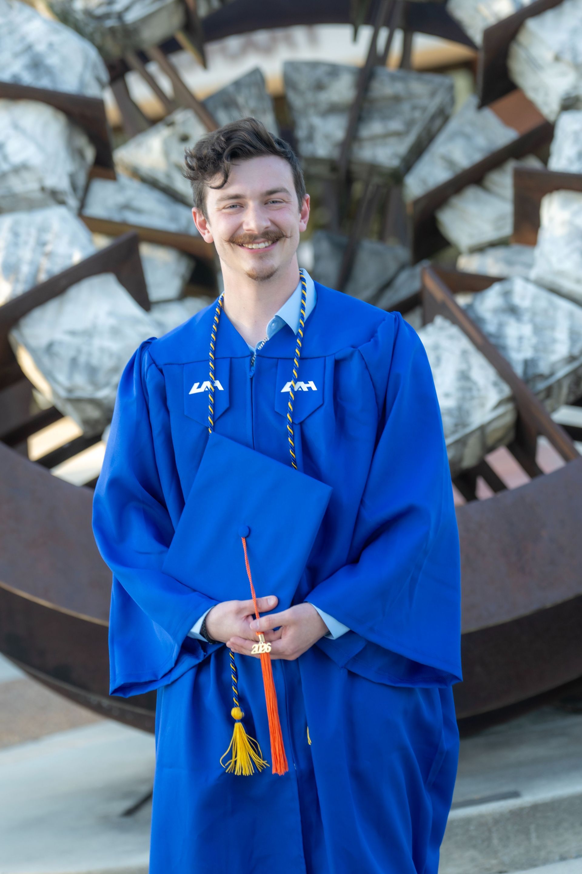 Person in a blue graduation gown holding a decorated item in front of a large stone sculpture.