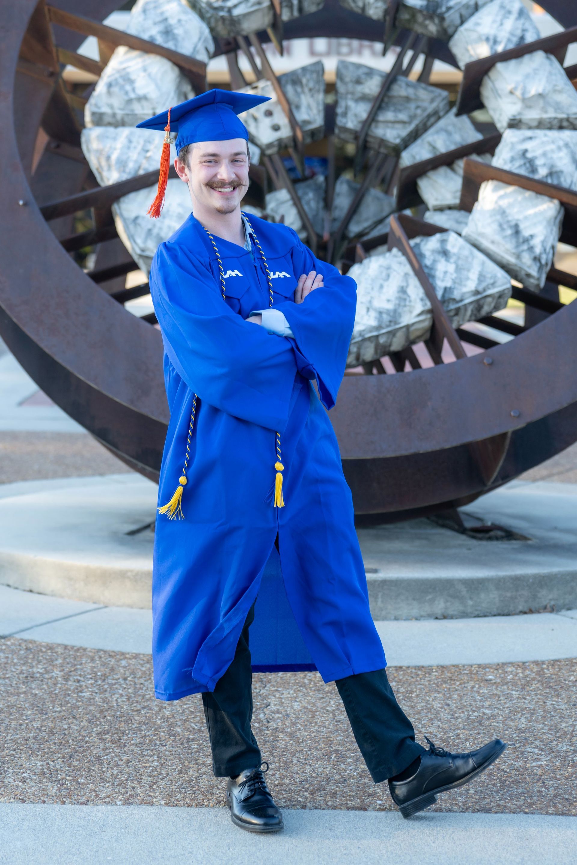 Person in blue graduation gown and cap posing in front of a large circular sculpture.