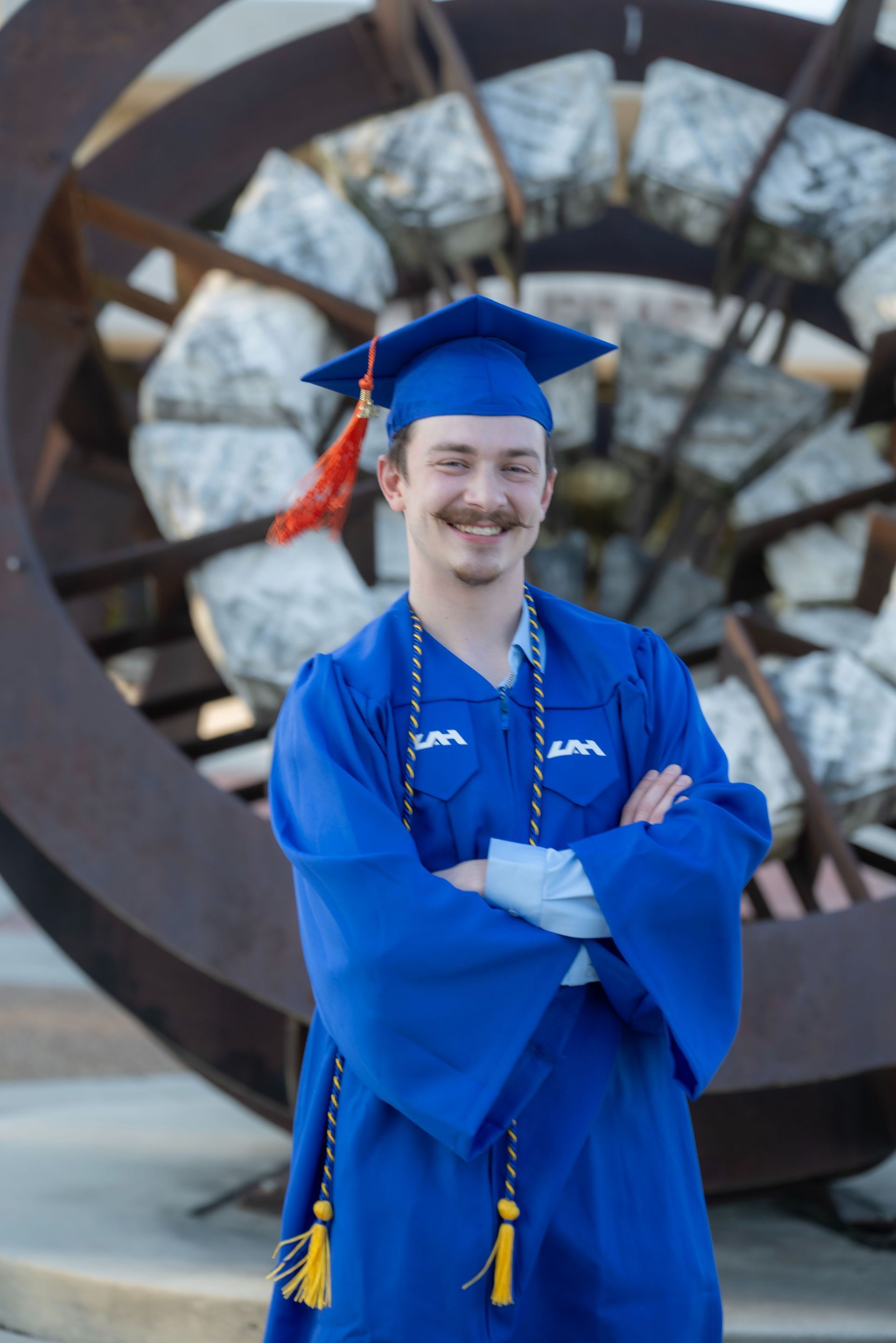 Graduate in blue cap and gown standing in front of a large metal sculpture, smiling with arms crossed
