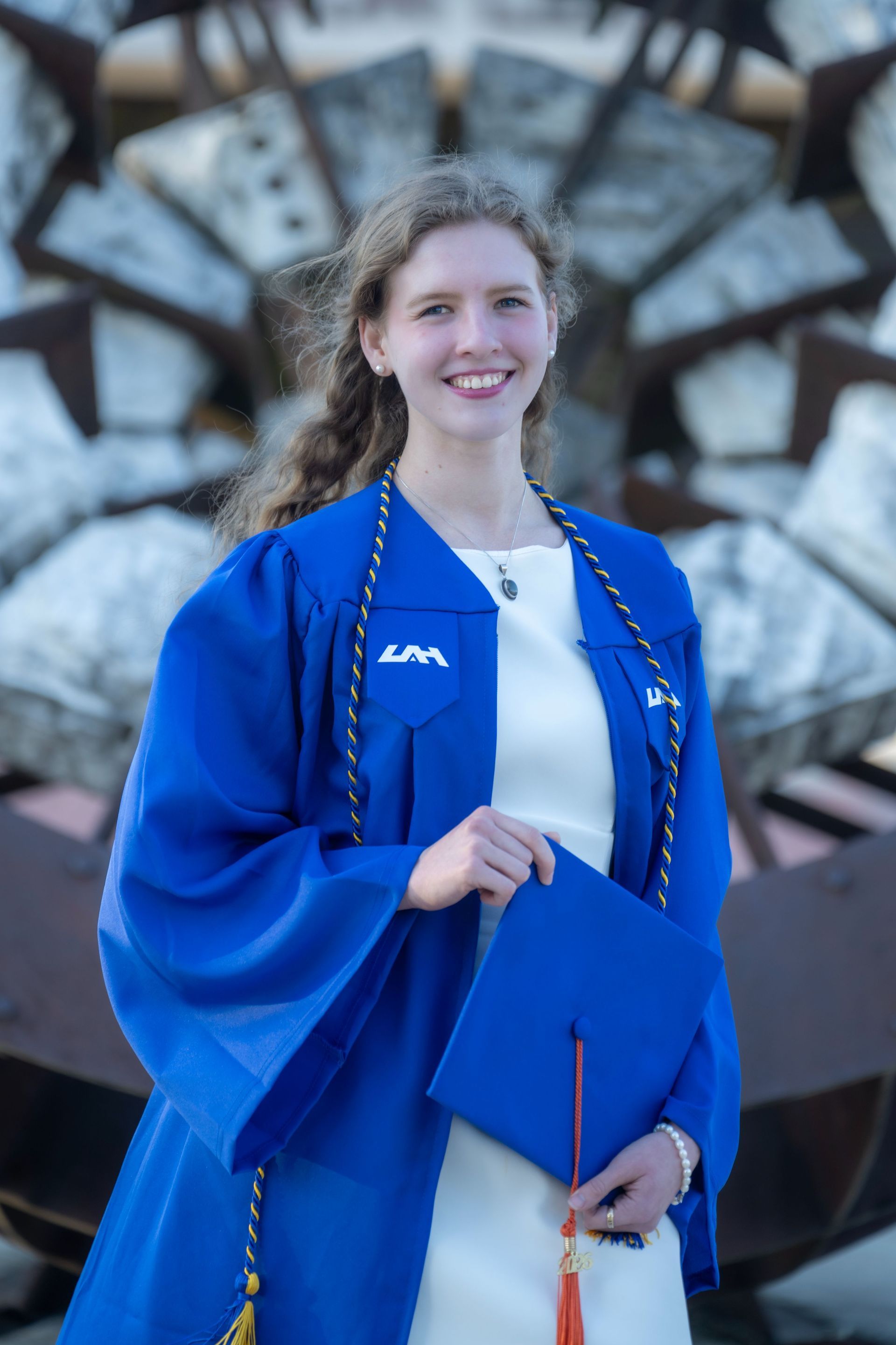 Person in blue graduation gown holding diploma, standing in front of a stone fountain.
