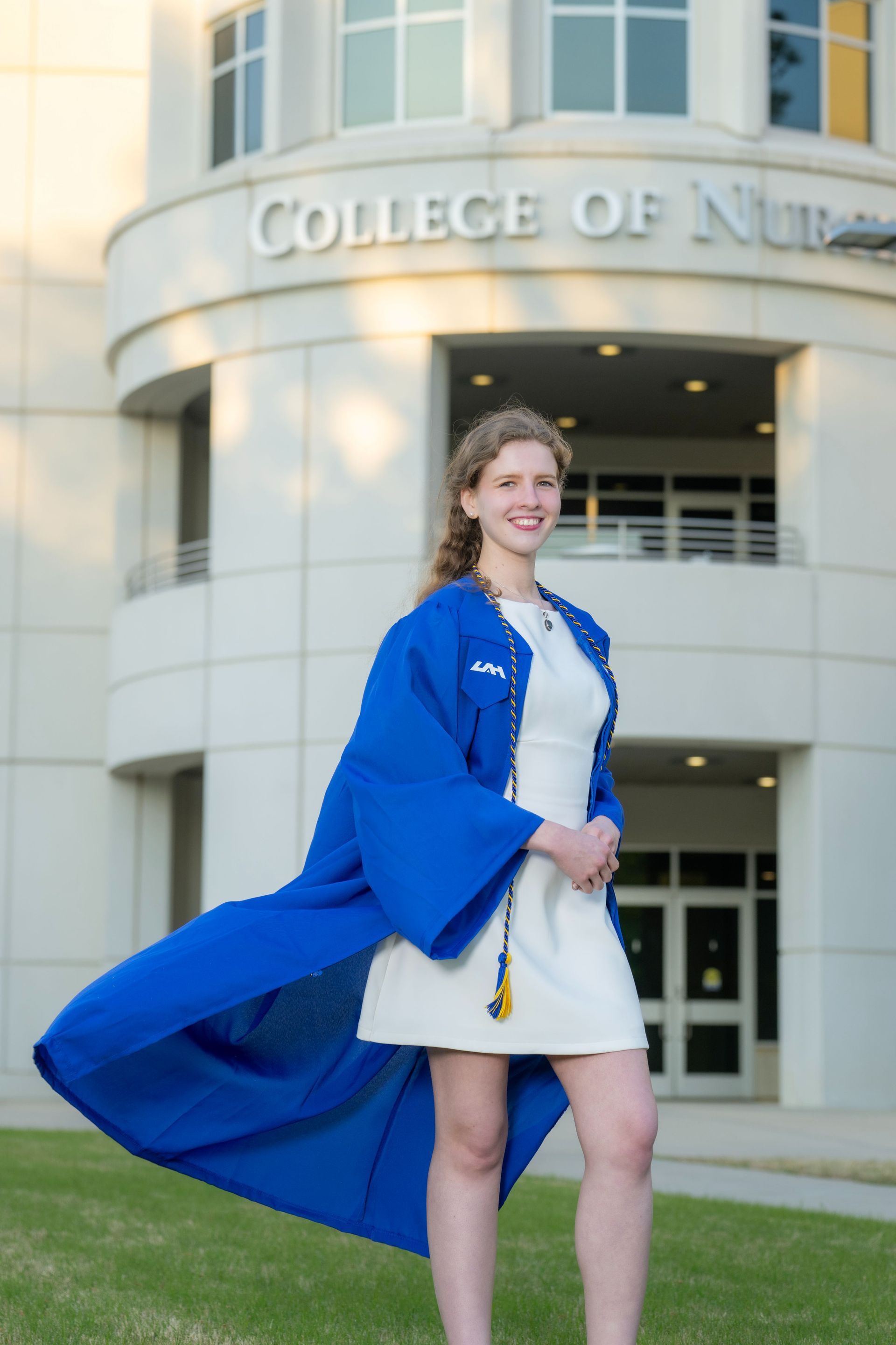 Graduate in blue coat and white dress standing outside a college building.