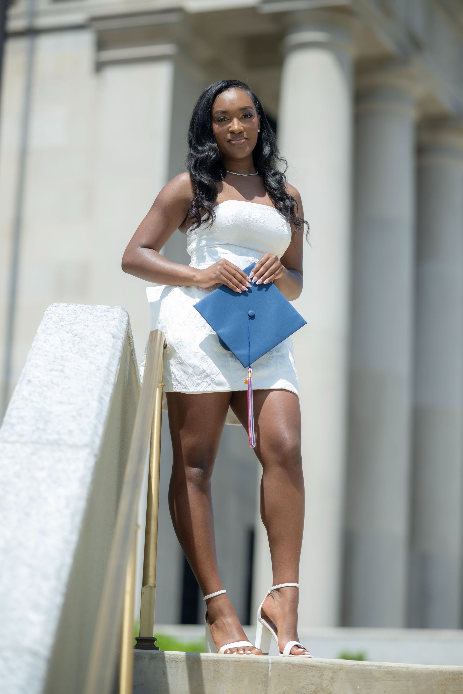 Woman in a white dress holding a blue clutch, standing on steps before tall columns.