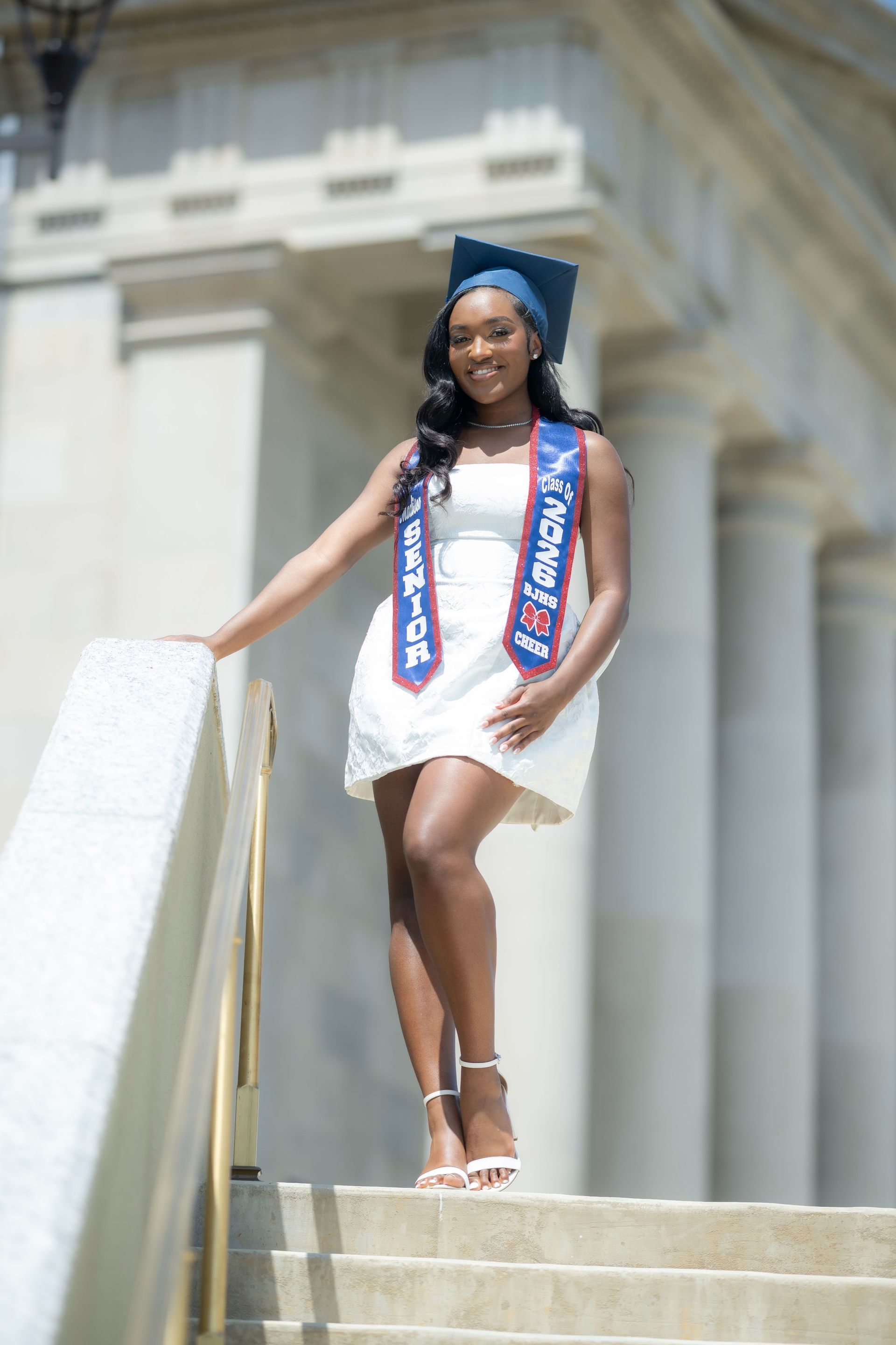 Woman in white dress and graduation cap standing on steps with a blue-red sash, smiling outdoors