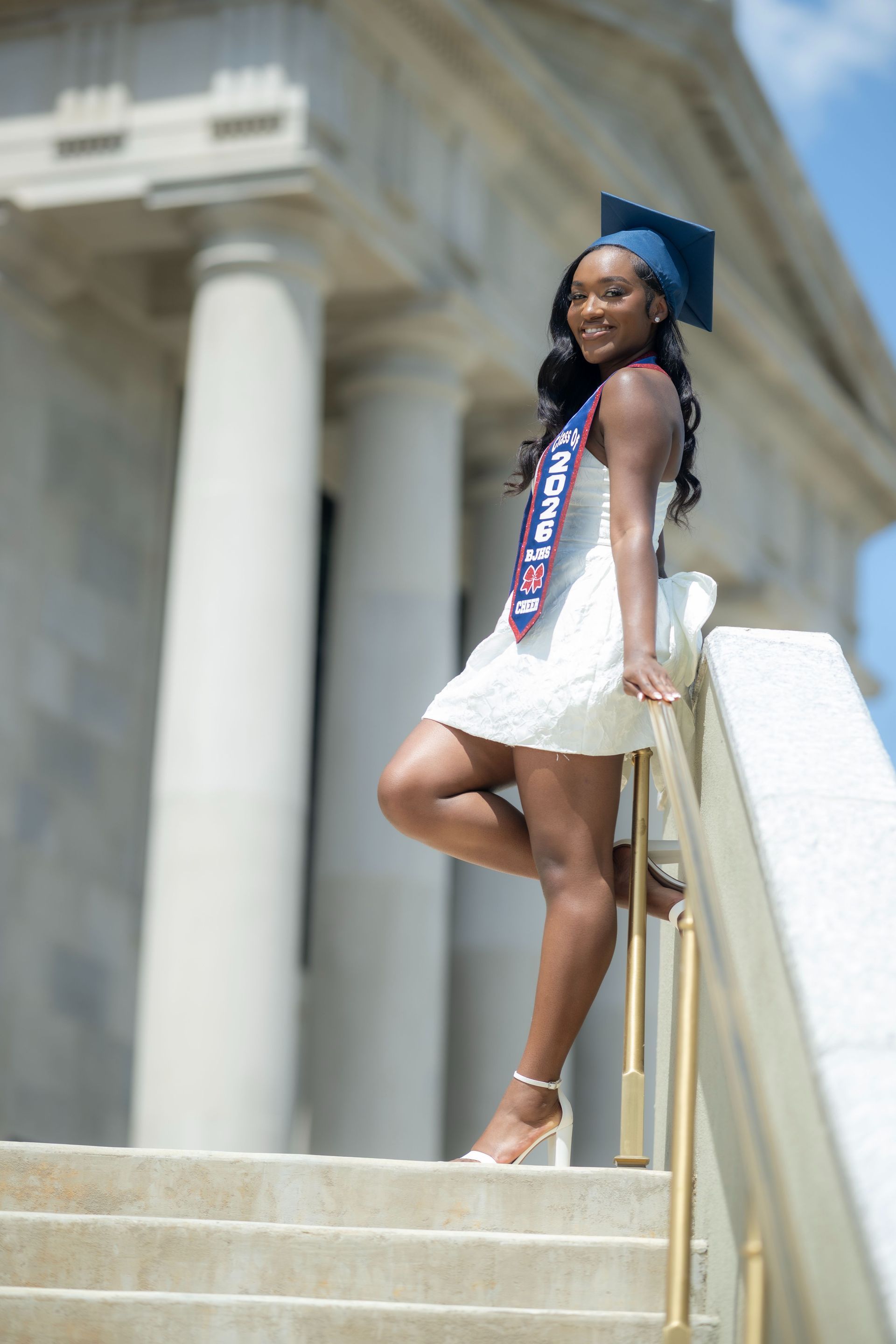 Graduate in white dress and blue cap posing on courthouse steps with diploma sash