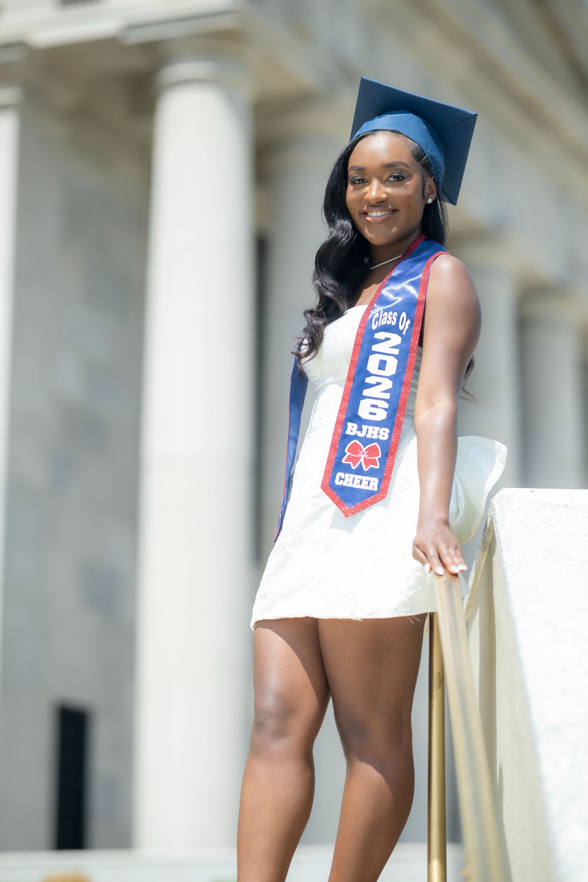 Graduate in blue cap and white dress standing outdoors on stone steps, wearing a blue sash.