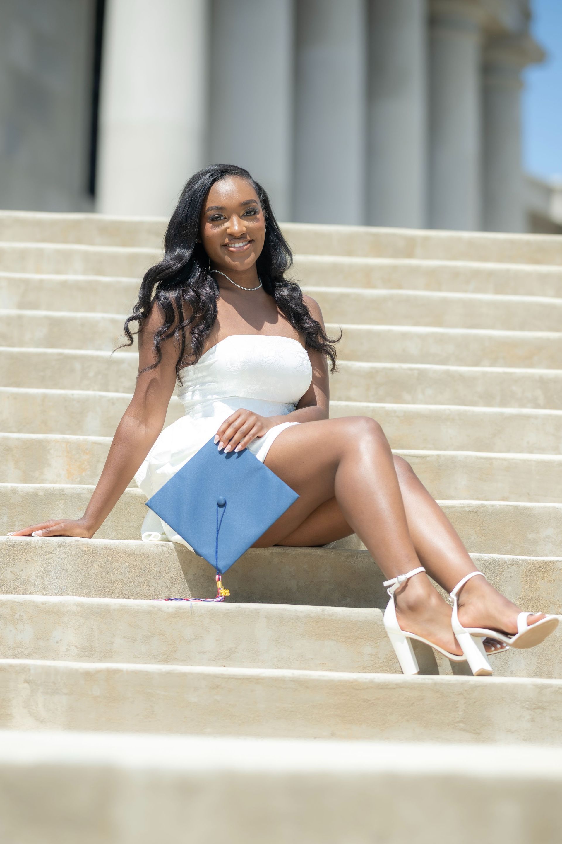 Woman in a white dress sitting on stone steps, holding a blue folder and smiling outdoors.