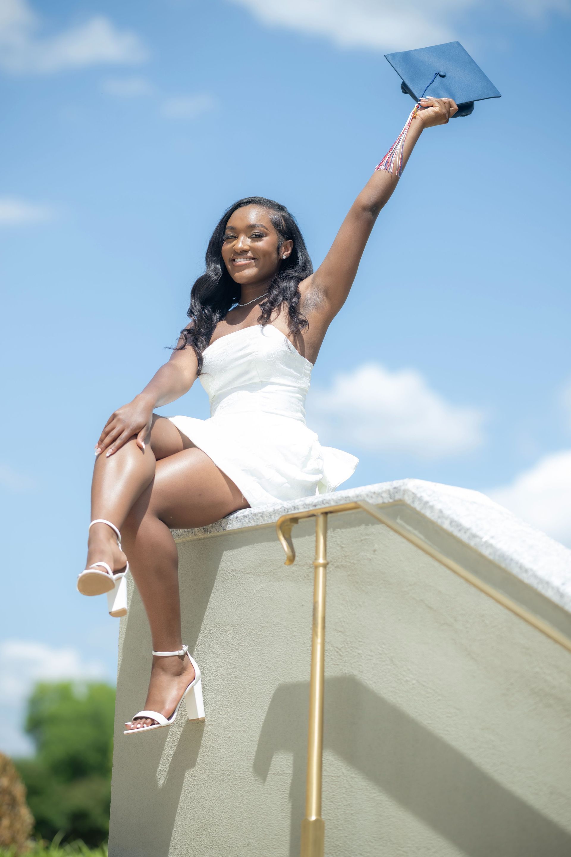 Woman in a white dress sitting on a ledge, smiling and holding a graduation cap against a blue sky.
