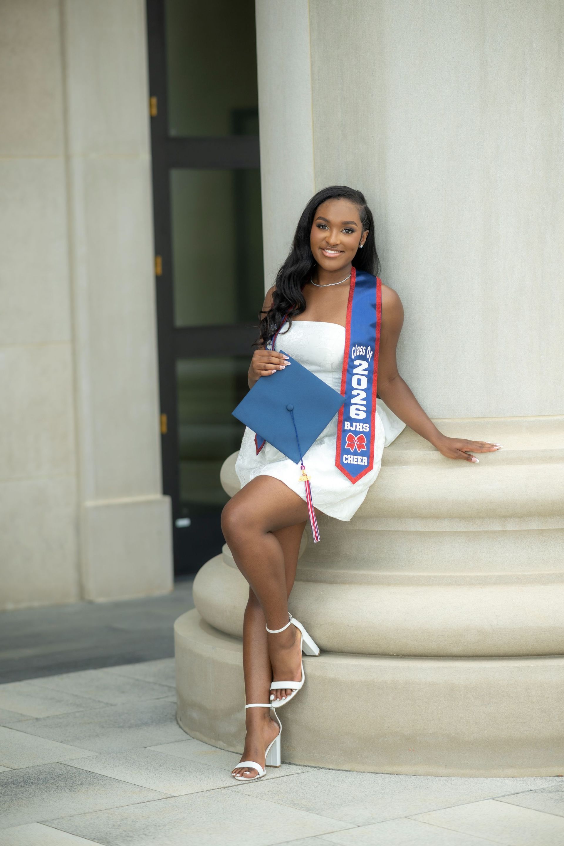 Woman in white dress and sash posing with a blue folder beside a column outdoors.