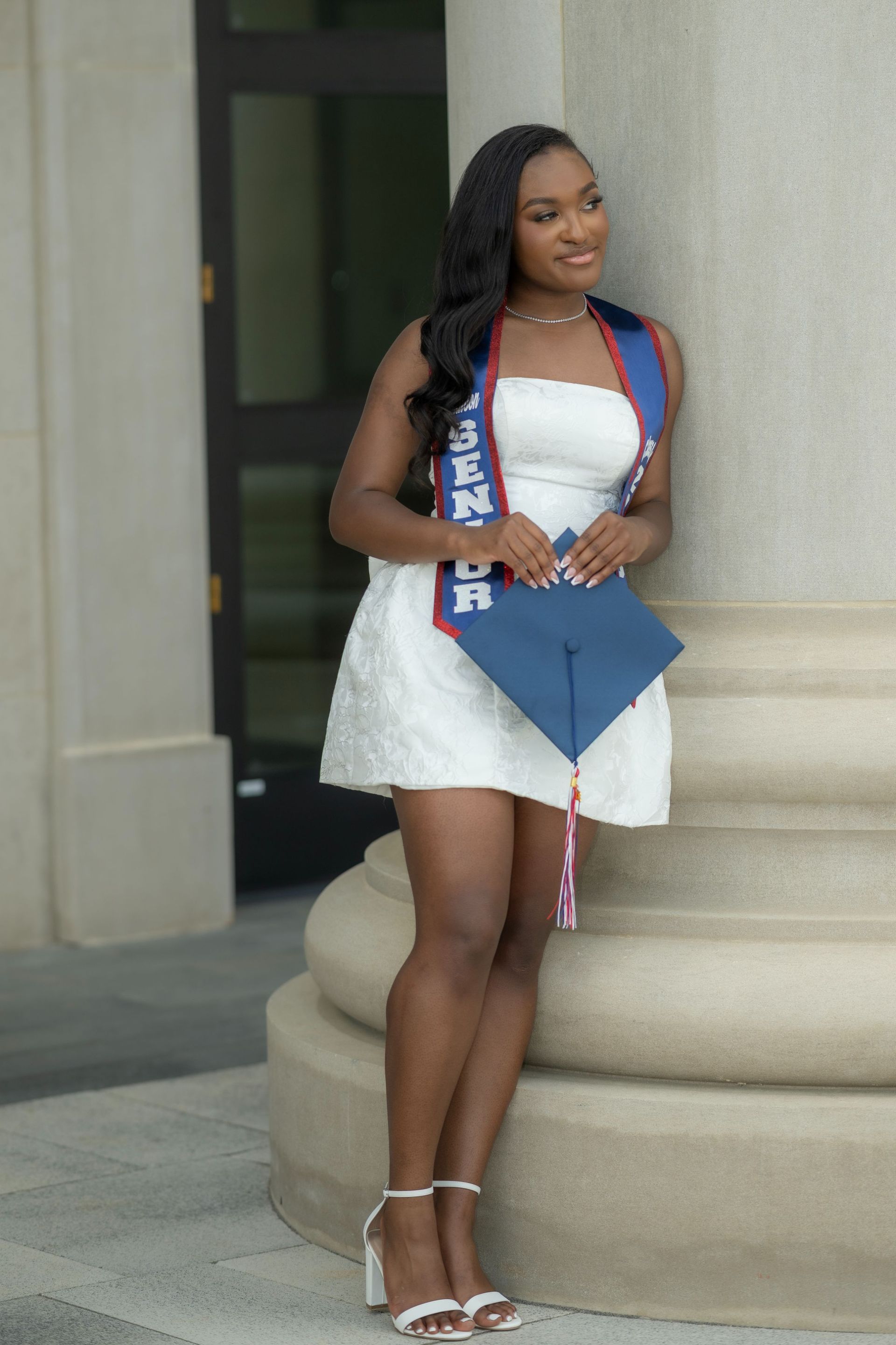 Person in a white dress holding a blue graduation cap, standing by a column.