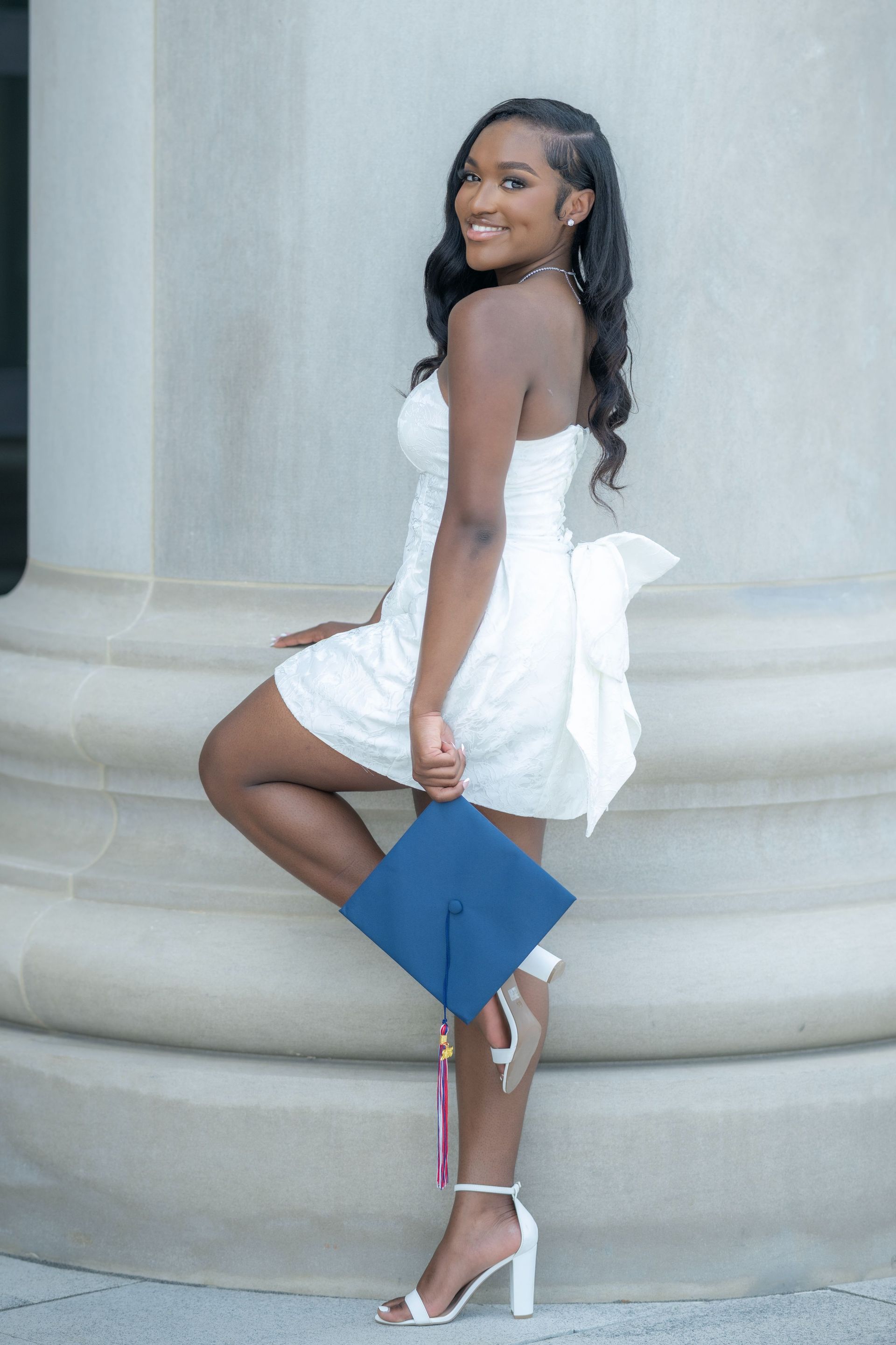 Woman in a white dress posing on steps, holding a blue clutch and smiling at the camera.
