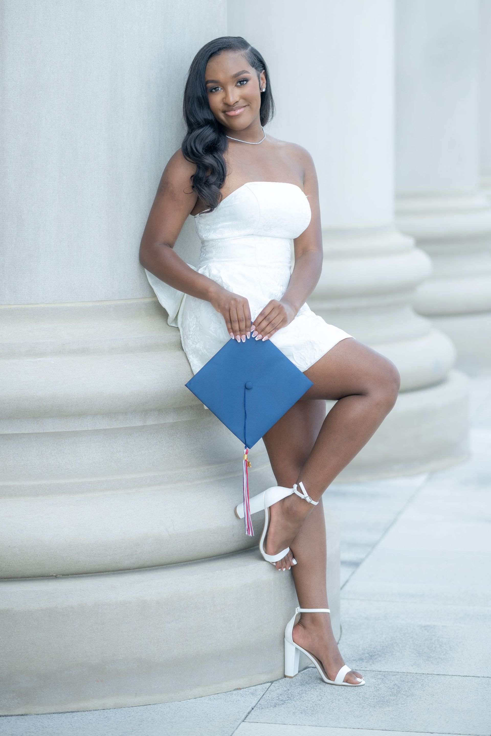 Woman in a white dress holding a blue graduation cap, seated by a large column outdoors.