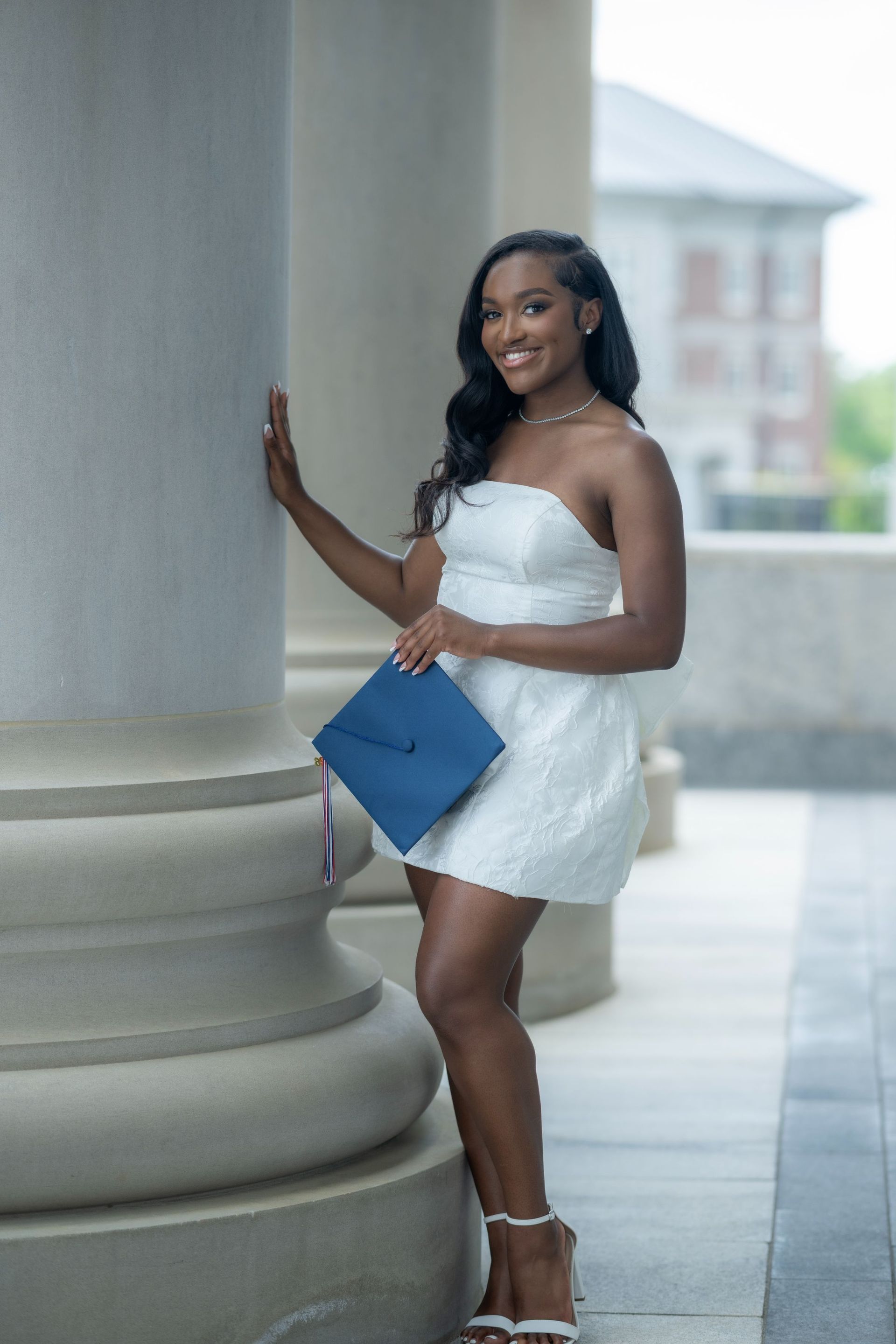 Woman in a white dress posing by a column, holding a blue clutch and smiling.