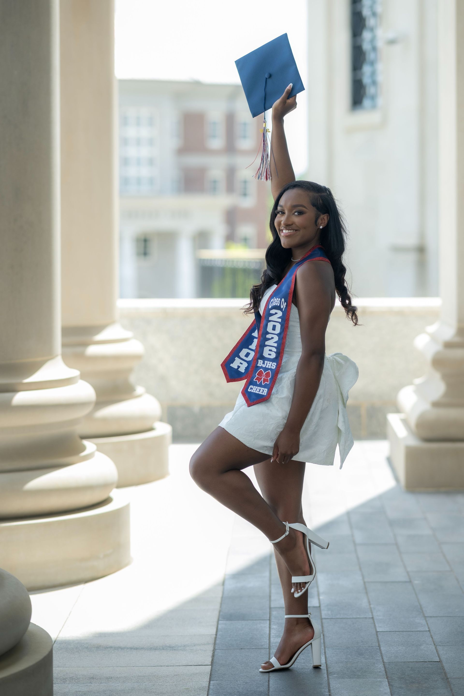 Graduate in white dress and heels posing outdoors with diploma raised