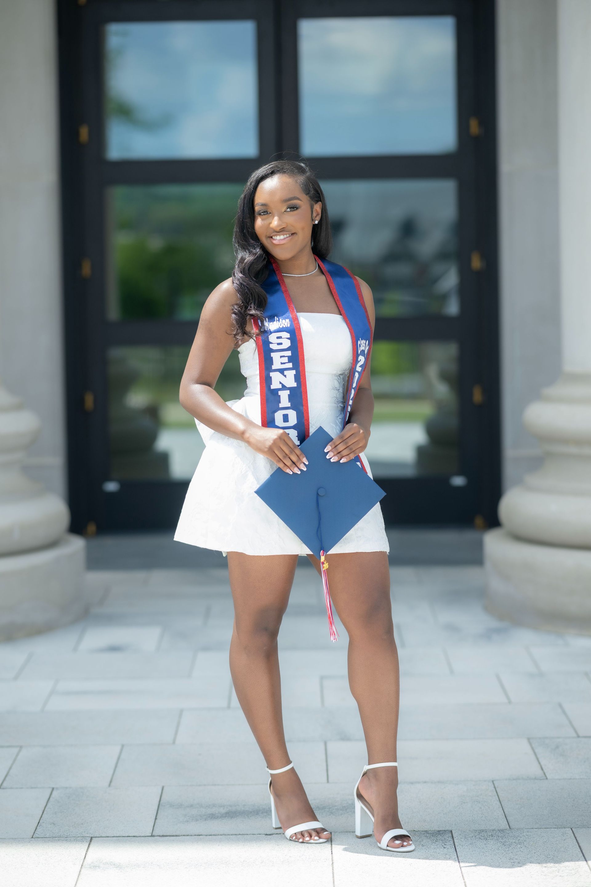 Person in white dress and heels holding a folder and sash, standing outside a building