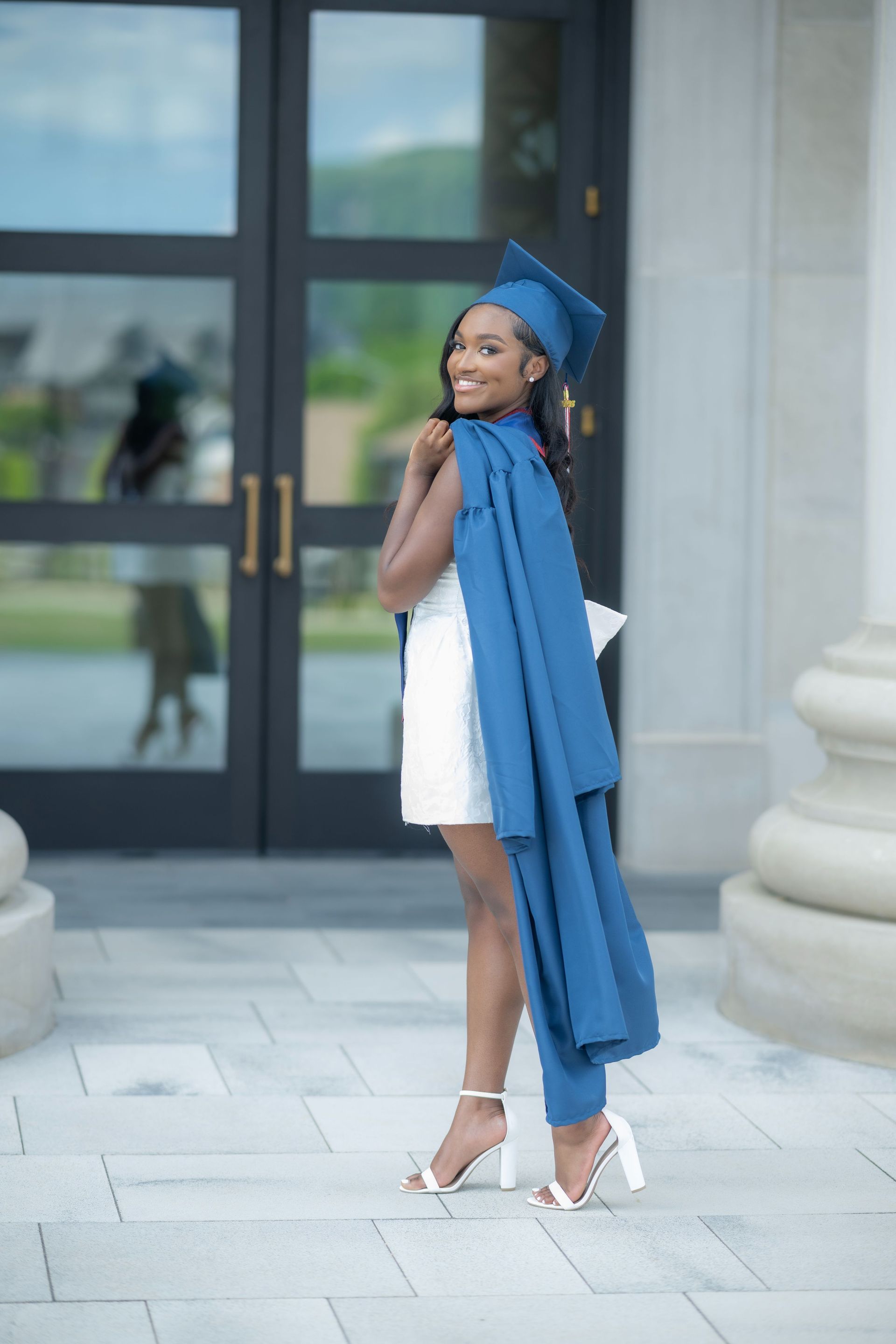 Person in a white dress and blue graduation cap and stole posing outdoors by glass doors.