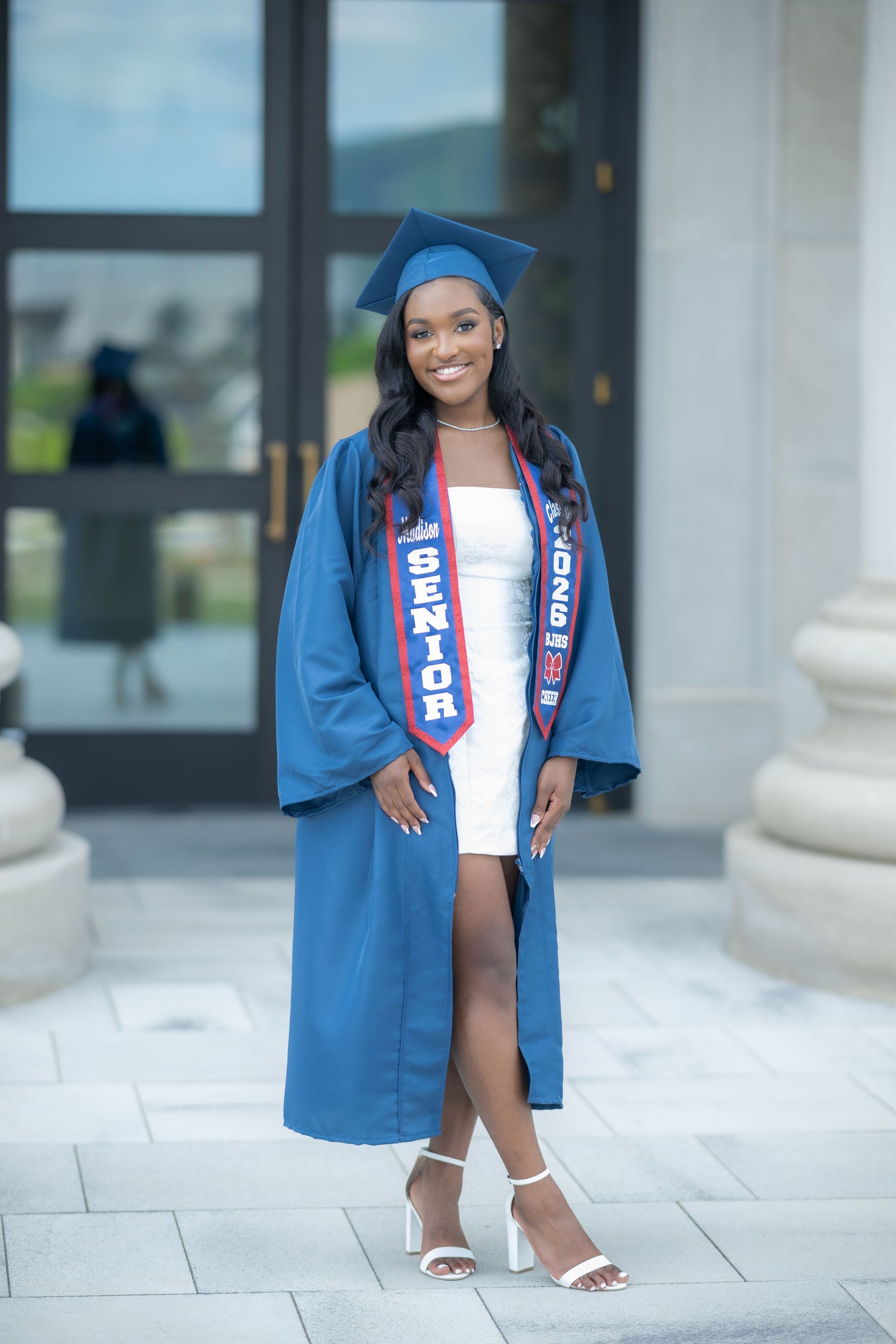 Graduate in blue cap and gown with pink dress, standing outside a building and smiling.