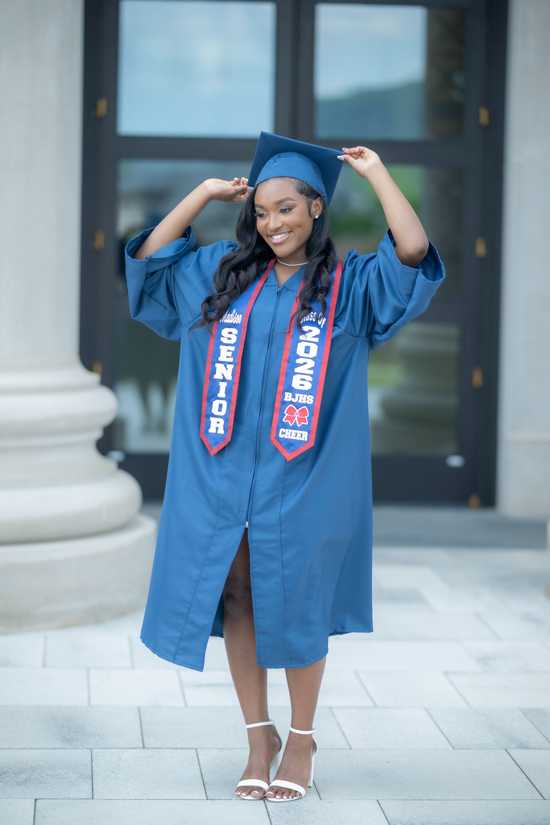 Graduate in blue cap and gown smiling outdoors in front of a building with columns