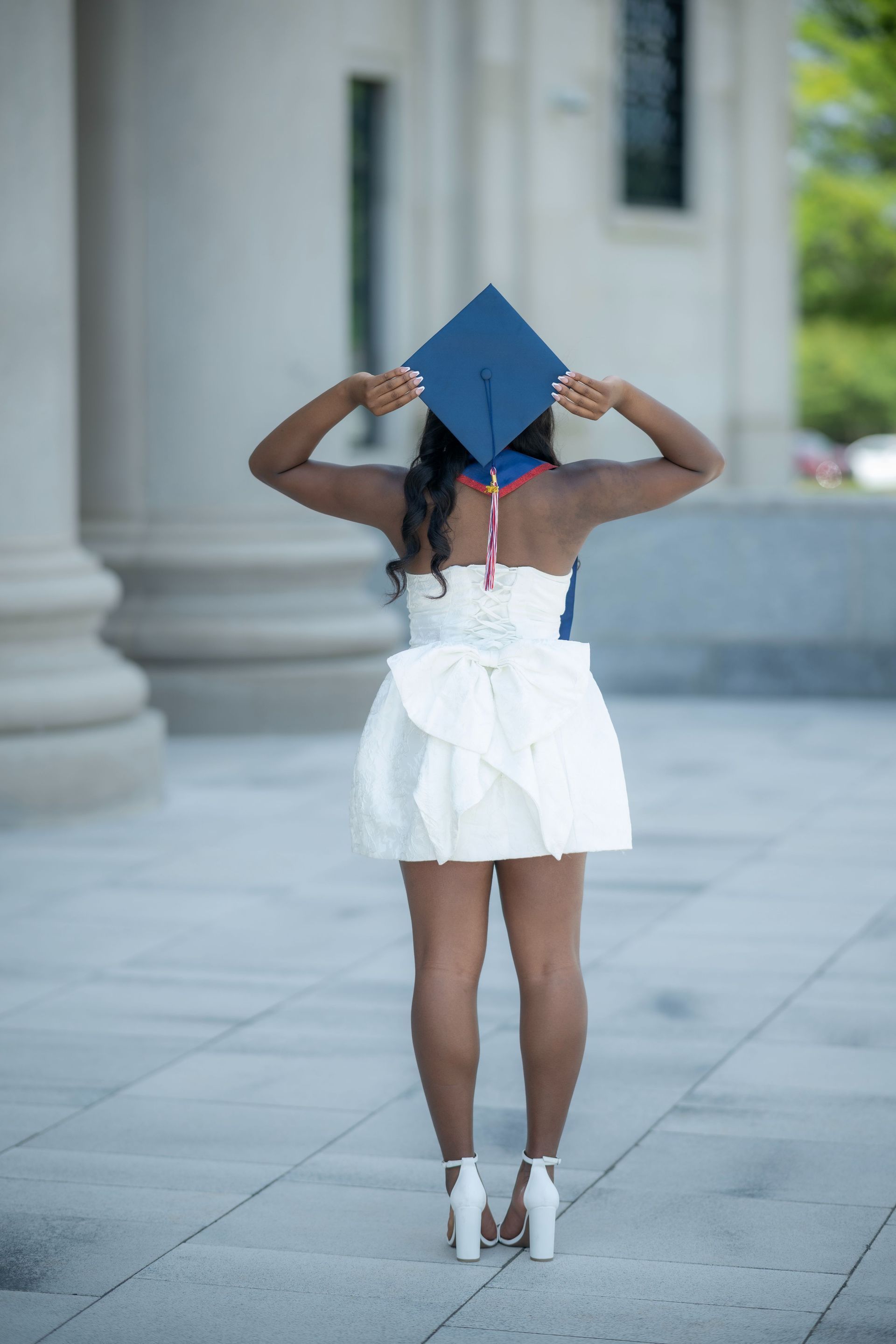 Person in a white dress and heels holds a blue graduation cap on a sidewalk.
