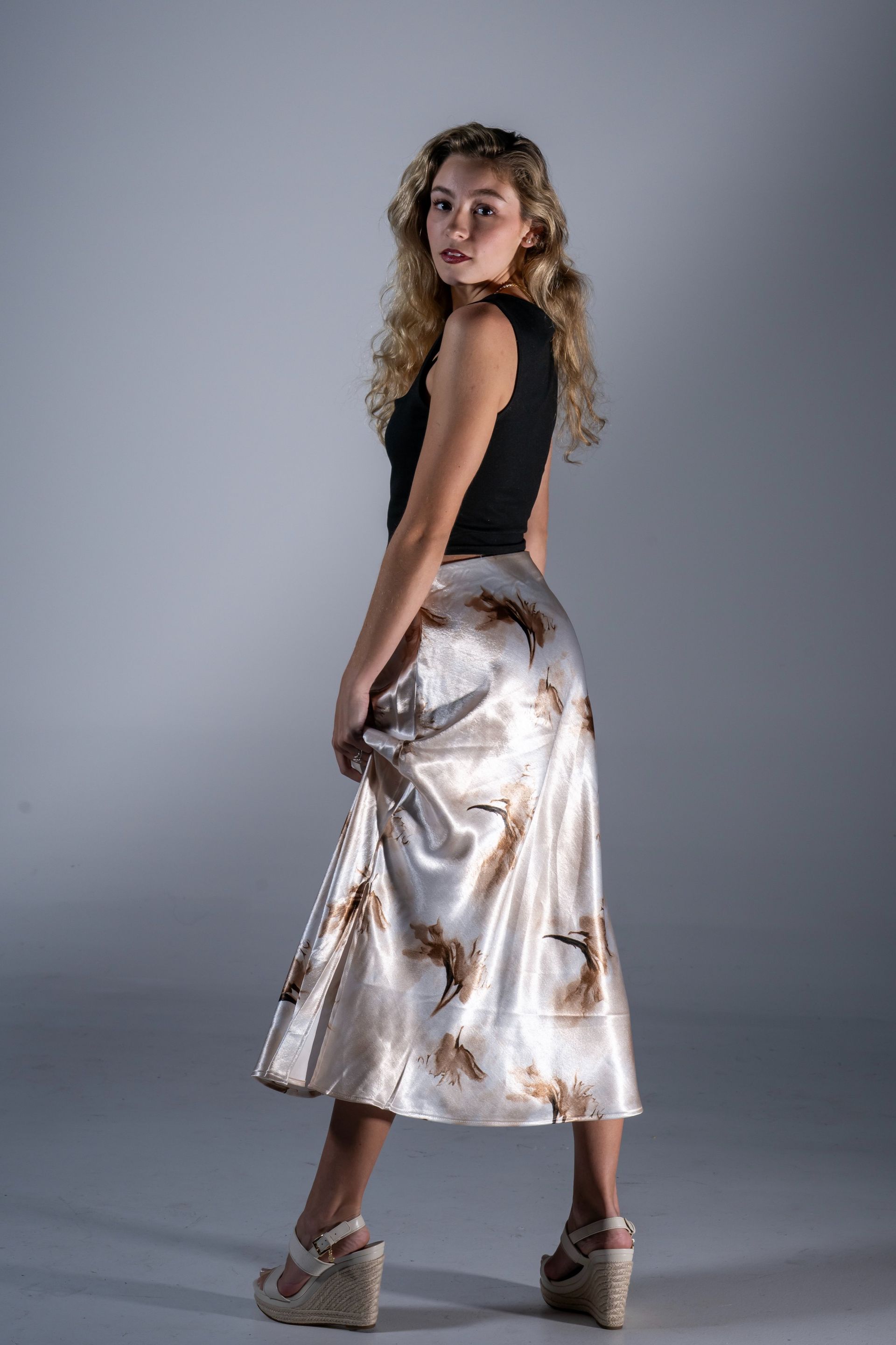 Person posing in a black top and metallic skirt against a gray studio backdrop, looking over their shoulder