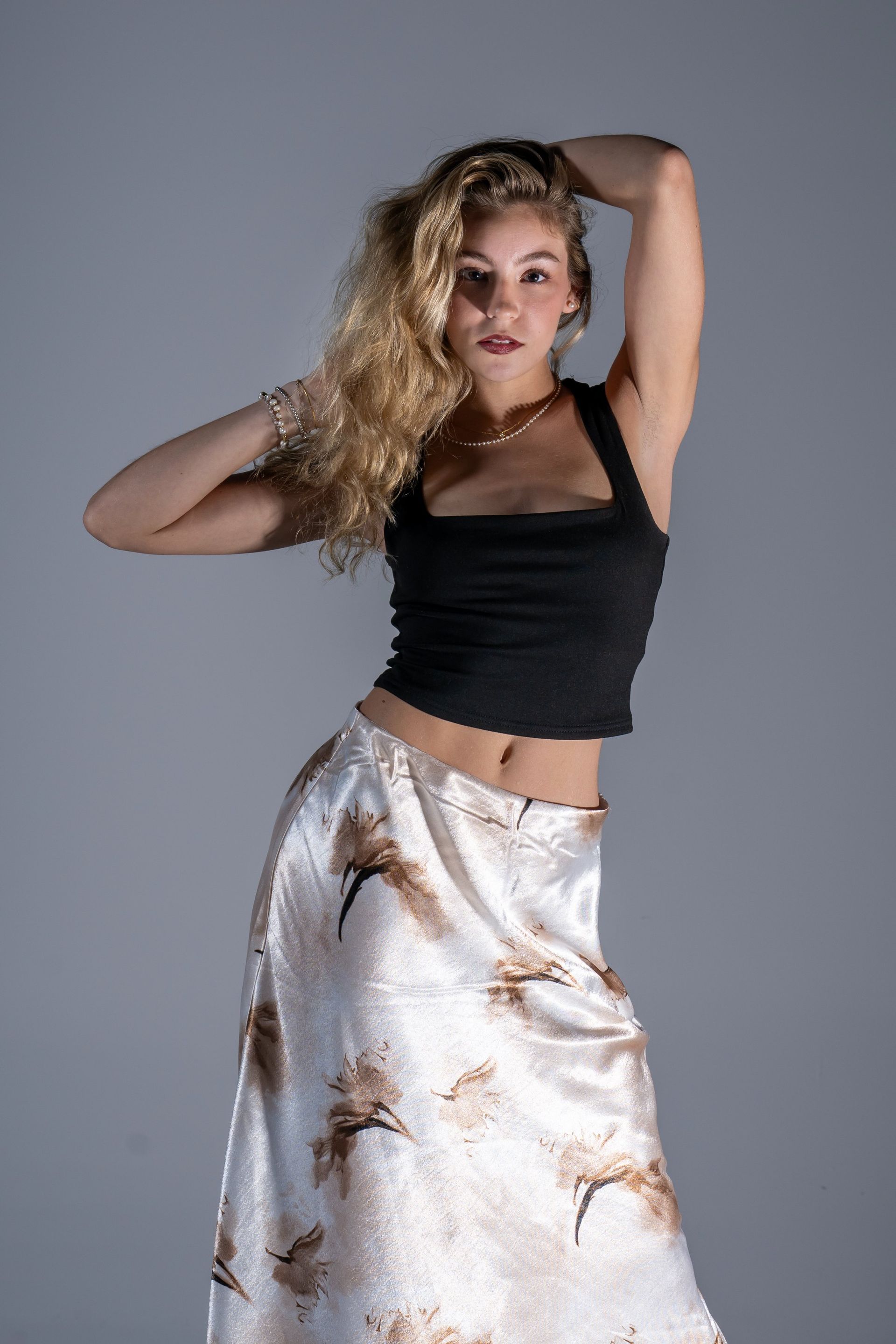 Woman posing in a black top and white satin skirt against a gray backdrop.