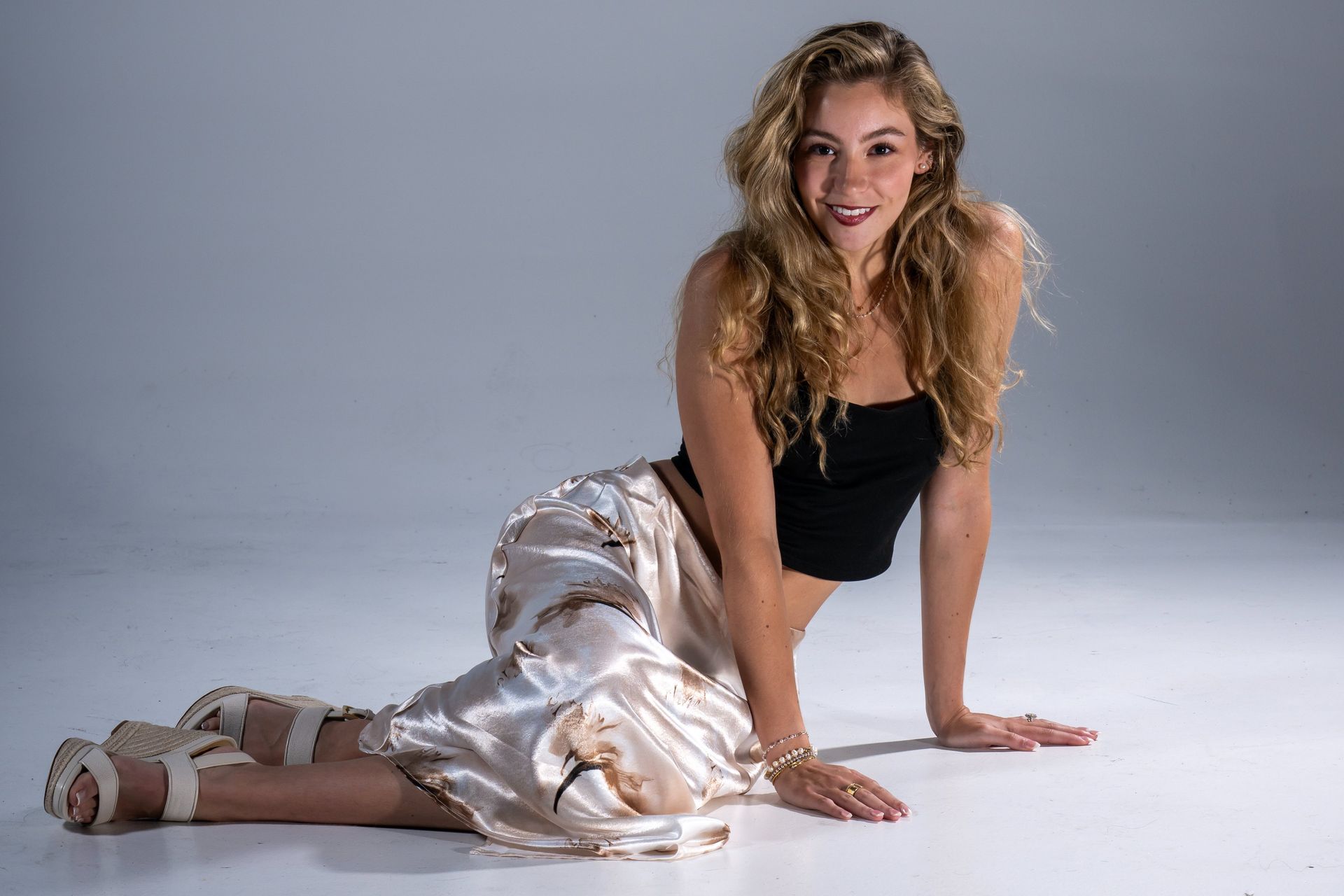 Woman kneeling on a studio floor, smiling in a black top and light satin pants.