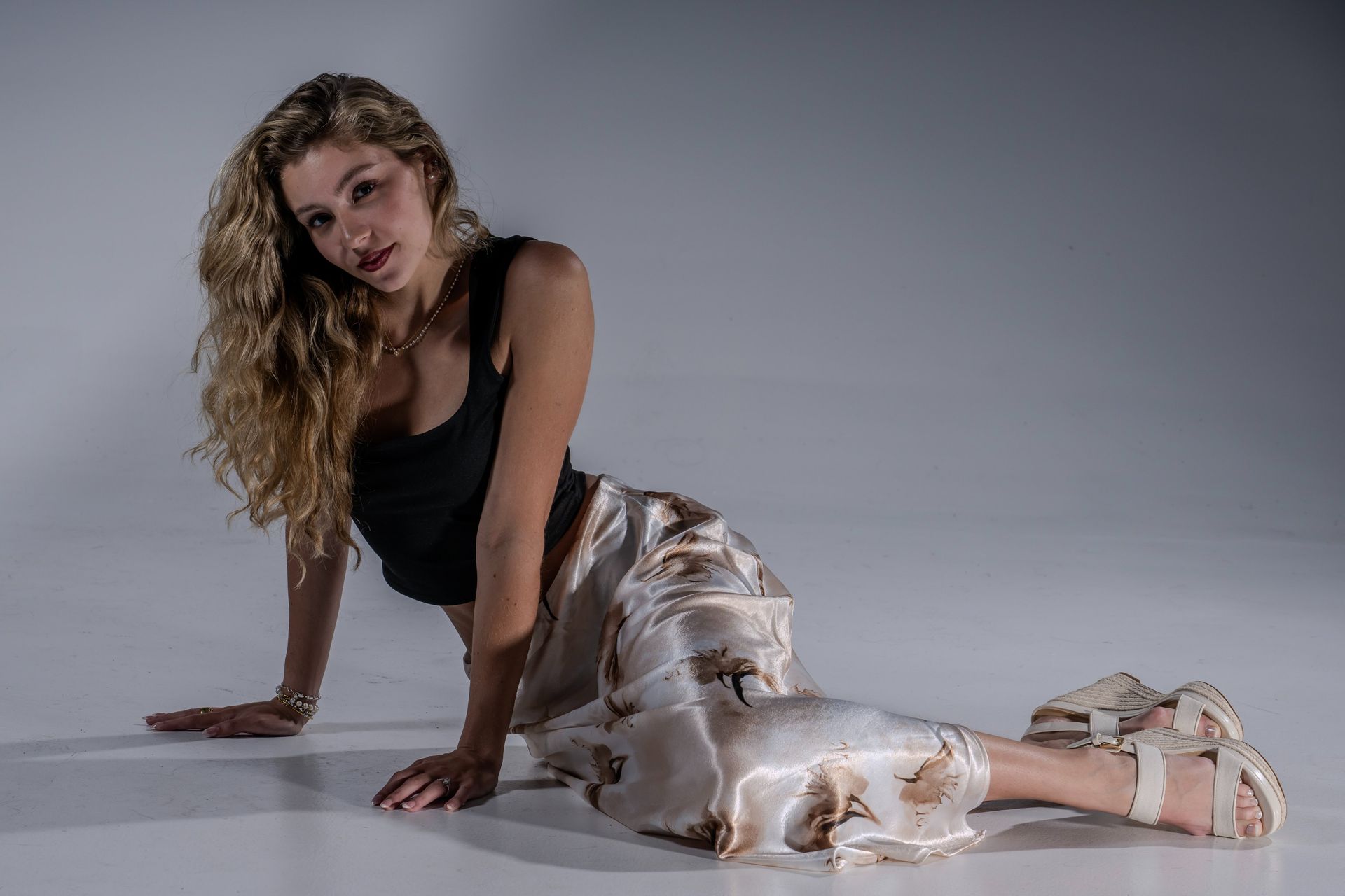 Woman posing seated on a studio floor in a black top and patterned pants, looking at the camera