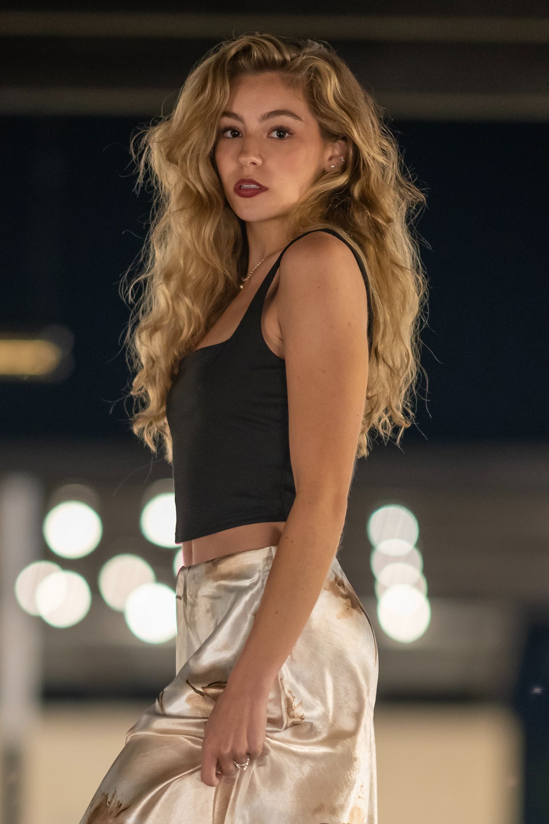 Woman in a black top and metallic skirt posing at night with blurred city lights behind her