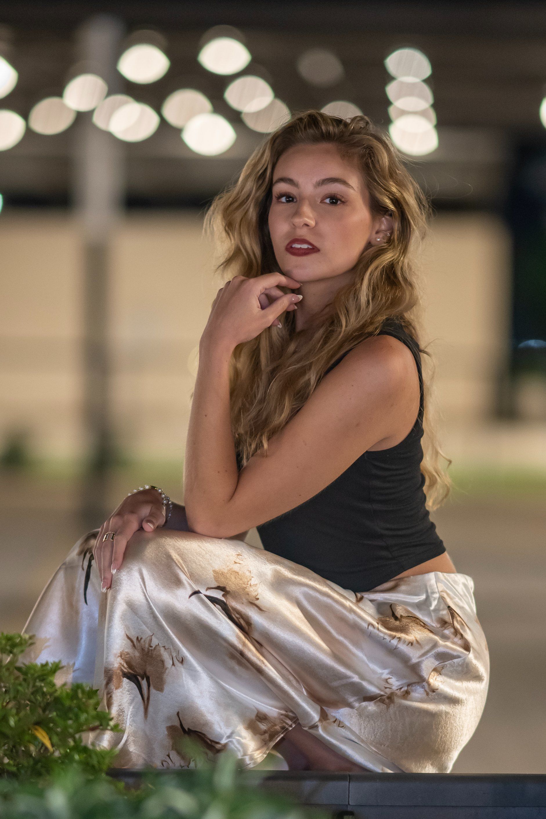 Woman in a black top and metallic skirt posing outdoors with blurred lights in the background