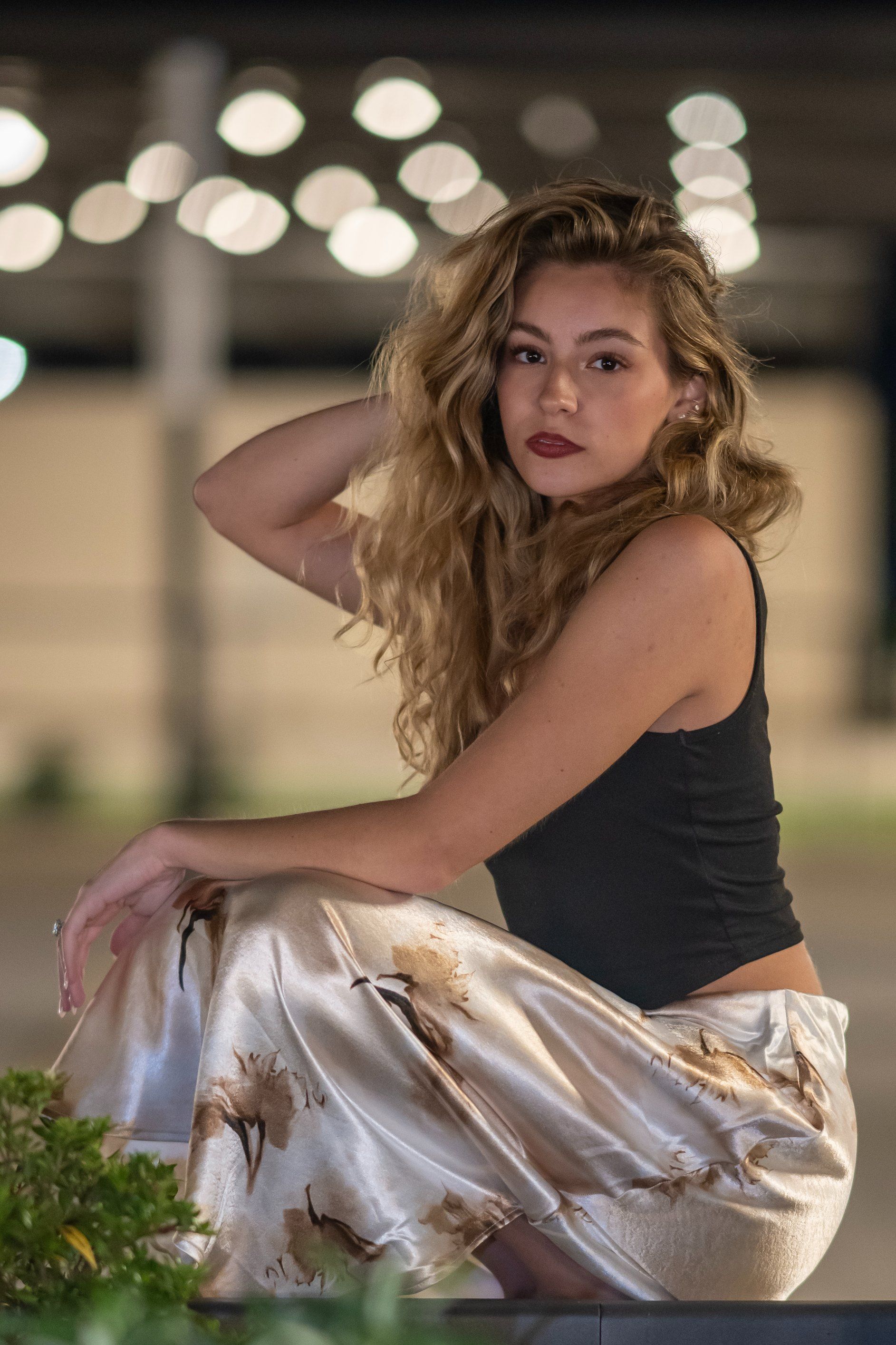 Woman posing outdoors at night in a black top and shiny gold pants, with blurred lights in the background