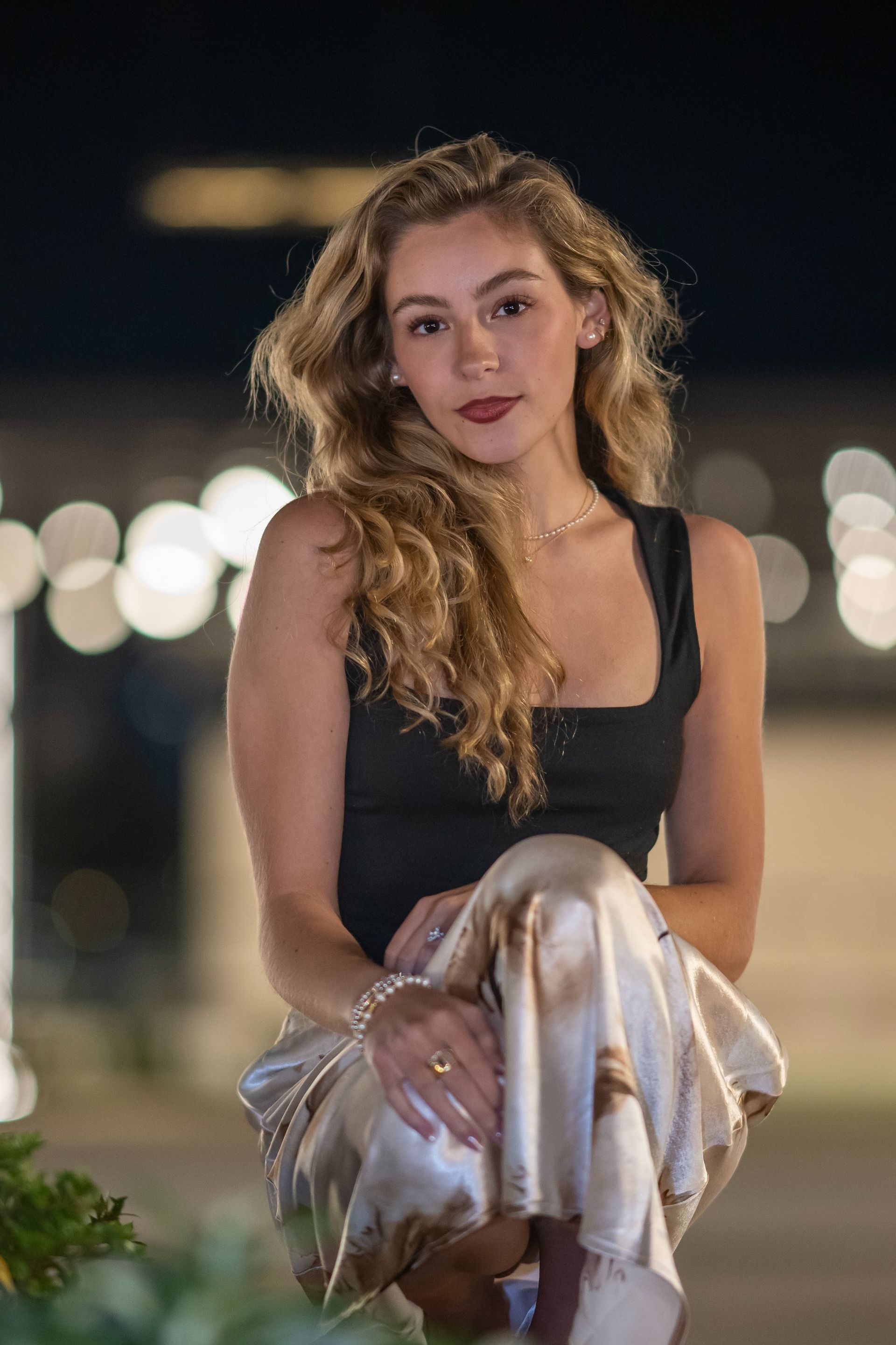 Woman in a black top holding a white jacket, seated outdoors at night with blurred city lights behind her.